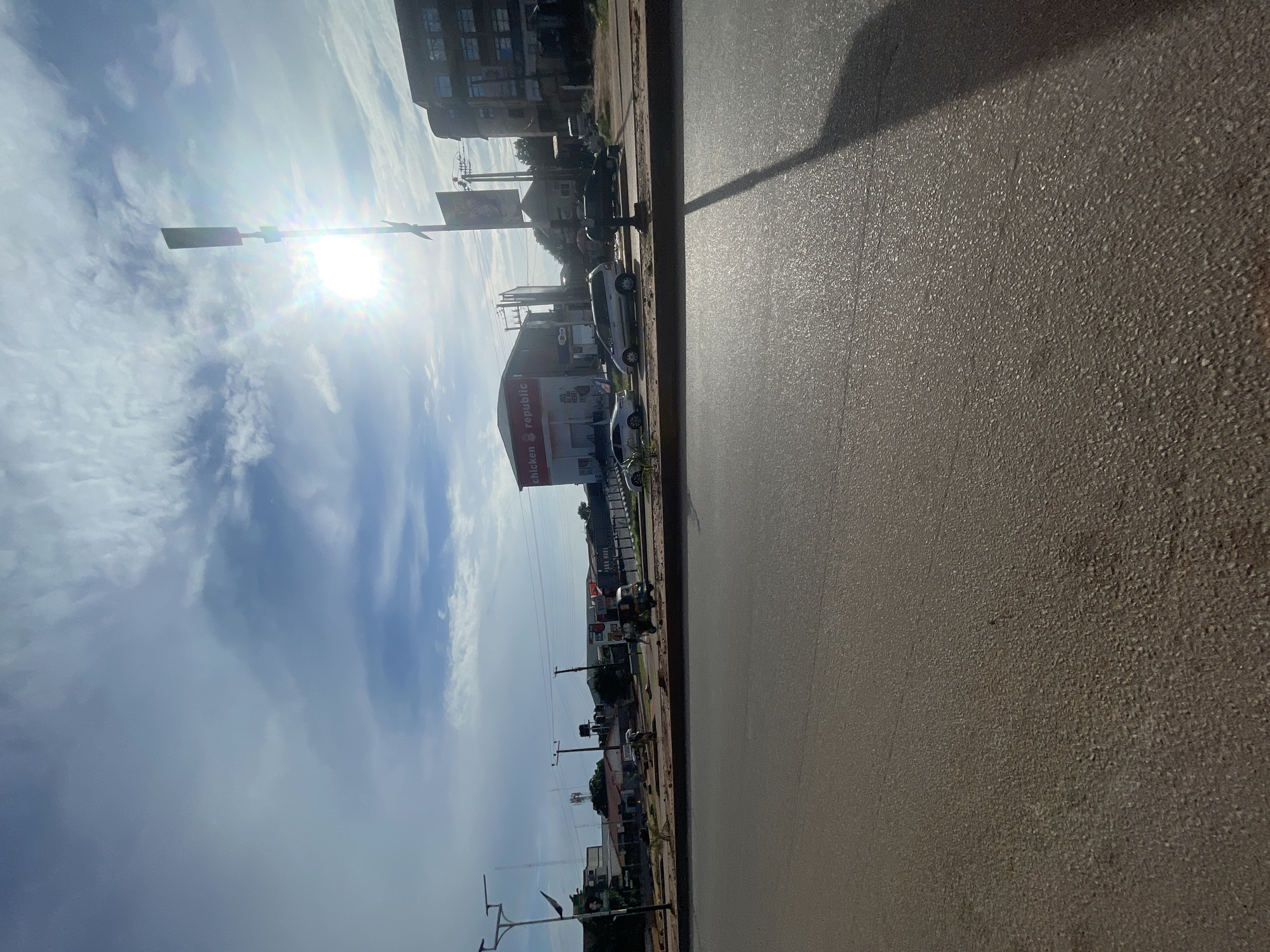 The image depicts a street scene in Jos, Nigeria, under a partly cloudy sky with the sun shining brightly. In the foreground, a paved road occupies the lower half of the frame, with a shadow of a street light extending across it from the right. Beyond the road, a median strip separates it from the opposite side of the street.
On the opposite side of the street, a row of buildings and businesses are visible. One prominent building displays the signage "CHICKEN REPUBLIC" in red letters. Several cars are parked or driving along this side of the road. A three-wheeled vehicle, commonly known as an "okada" or Keke Napep, is also present, with a person on it. In the background, more buildings, trees, and utility poles can be seen. The overall impression is of a commercial area in an urban environment in Jos.