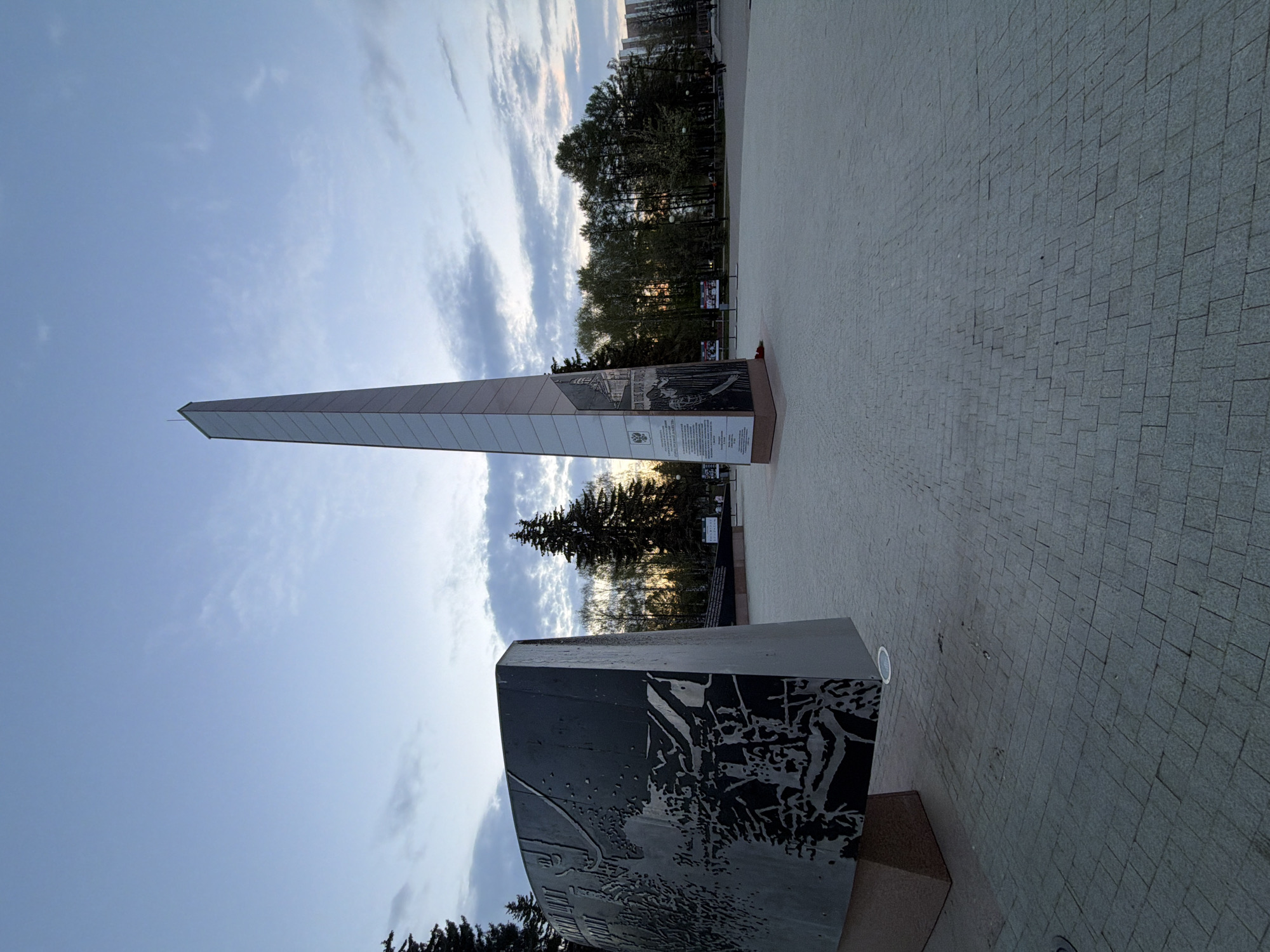 The image depicts a monument in Izhevsk, Russia. It consists of a tall, thin obelisk and two shorter, wider rectangular structures with engraved surfaces. The sky is partly cloudy, suggesting either sunrise or sunset. Trees are visible in the background. The ground is paved with small, grey square tiles. There are no people visible in the image.