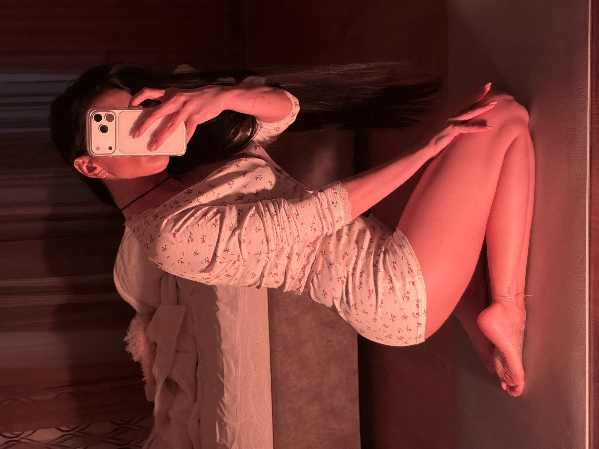 A woman is lying on a bed taking a selfie with her phone. She is wearing a floral patterned pajama set. Her long dark hair is spread out. The location cannot be determined from the image, and is not Svkh Voronezhskiy, Russia.