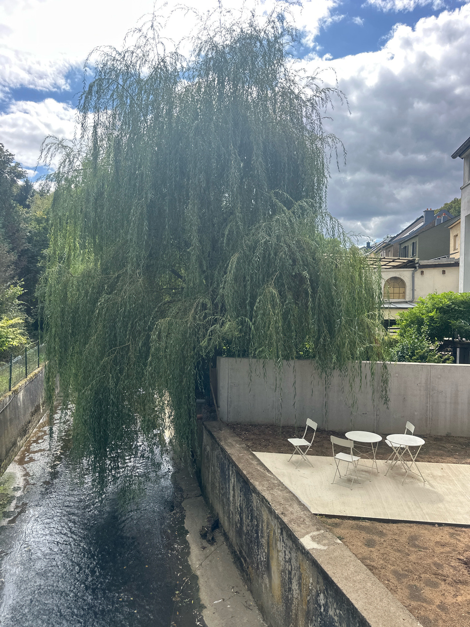The image depicts a scene in Bridel, Luxembourg, featuring a narrow canal with a concrete embankment on the right side. Overhanging the canal and the embankment is a large weeping willow tree with its branches reaching towards the water. On the concrete embankment, a light-colored paved area hosts a small outdoor seating arrangement consisting of two white, foldable bistro tables and three matching white, foldable chairs. The tables are round, and one has a single chair positioned beside it, while the other has two chairs. Behind the paved area, a concrete wall separates the embankment from a residential building with a sloping roofline and windows visible. The sky above is partly cloudy with a mix of white and dark gray clouds. The canal water appears dark and somewhat murky.