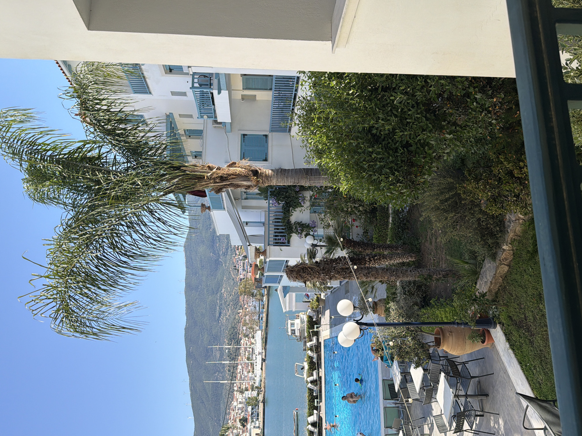 This vibrant, sunny image captures a scenic view of Poros, Greece, from an elevated perspective, likely a balcony or terrace. In the foreground, a white architectural element, possibly a balcony railing or wall, frames the top and right side of the image. A large palm tree with lush green fronds occupies the left side, partially obscuring the view but adding a tropical touch.
The midground features a resort-like setting with white buildings characterized by traditional blue balconies and shutters, indicative of Greek island architecture. Lush green bushes and trees are interspersed between the buildings and surrounding a sparkling blue swimming pool. Several people are visible in the pool, enjoying a swim and cooling off. Adjacent to the pool, an outdoor seating area with tables and chairs suggests a restaurant or lounge. Decorative elements like large terracotta pots and lampposts with globe lights add to the ambiance.
Beyond the pool and buildings, a narrow channel of calm blue sea stretches into the distance, with numerous boats moored along its edges, forming a lively harbor or marina. On the opposite side of the channel, a picturesque town with white and terracotta-roofed buildings climbs up the gentle slopes of a large, verdant mountain. Further in the background, more mountains rise, dotted with a few wind turbines.
The weather is clear and sunny, with a bright blue sky, indicating daytime, likely mid-morning or afternoon. The overall atmosphere is one of leisure, vacation, and tranquility in a beautiful Mediterranean coastal setting. No discernible text is visible in the image.
