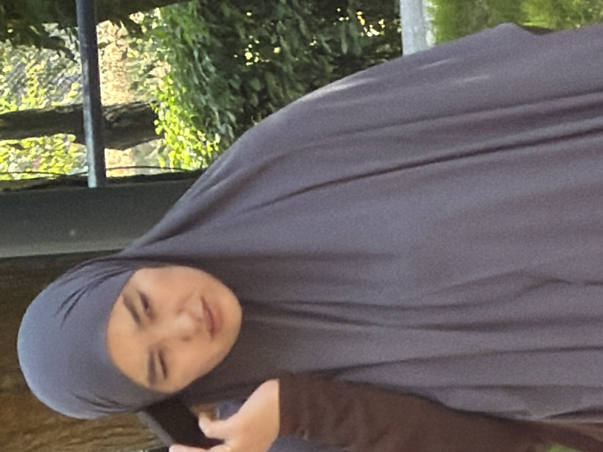 A medium shot captures a woman wearing a dark grey hijab, her face partially visible as she holds a phone to her ear. She appears to be looking down and to her left, with a subtle frown. Her left hand is raised and positioned near her face, touching the phone. She is wearing a dark brown long-sleeved shirt beneath the hijab. The background is blurred, showing greenery and trees, suggesting an outdoor urban setting, possibly a park or a street. The lighting is natural, indicating it is daytime.