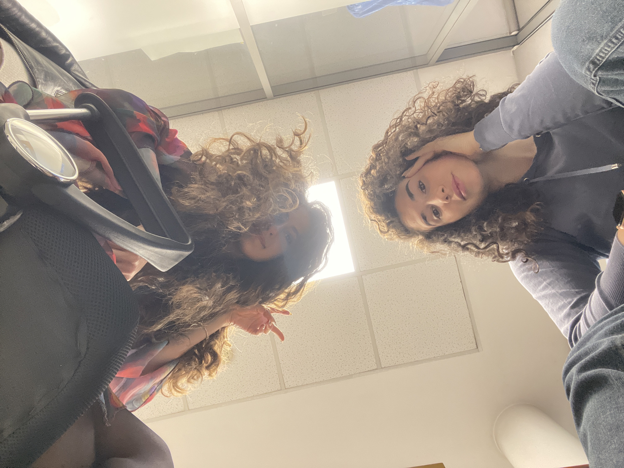 An extreme low-angle shot captures two young women with abundant curly brown hair looking down towards the camera. The woman on the left is partially obscured by a black chair with a silver armrest. She appears to be wearing a colorful, patterned top or dress, possibly layered under a black jacket. Her left hand is visible, partially raised as if in a gesture. The woman on the right wears a grey or dark blue hoodie and blue jeans. Her right hand is near her head, touching her hair, and she has a soft smile or smirk, with bright, engaging eyes.
The background consists of a white ceiling made of square acoustic tiles with visible seams, commonly found in office or institutional settings. Several bright fluorescent light fixtures are integrated into the ceiling, creating prominent illumination and some glare, especially in the upper center of the frame. The overall lighting suggests an indoor environment, likely well-lit by artificial means, possibly during the day. The perspective implies the photo was taken from directly below the subjects, possibly while they were looking down at the camera, giving the impression of the viewer being observed from above. No text is visible in the image. The location context is Sofia, Bulgaria.