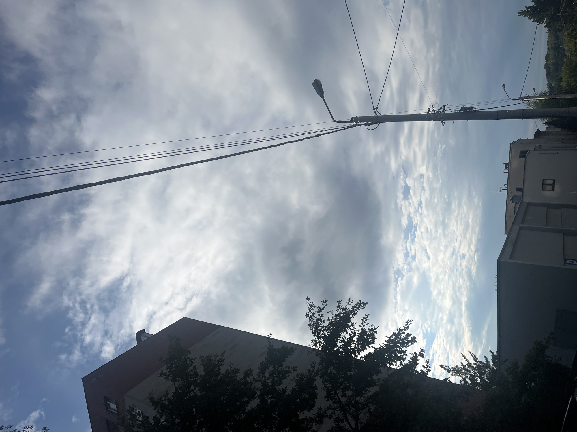 A low-angle shot shows a building with a red top against a cloudy sky. In the foreground, power lines stretch diagonally across the frame, with a lamppost on the right. The building is dark on the left side, with a tree casting a shadow. The sky is filled with a mix of gray and white clouds, with patches of blue visible. Several other buildings are visible in the background, and lush green trees form the horizon on the far right.