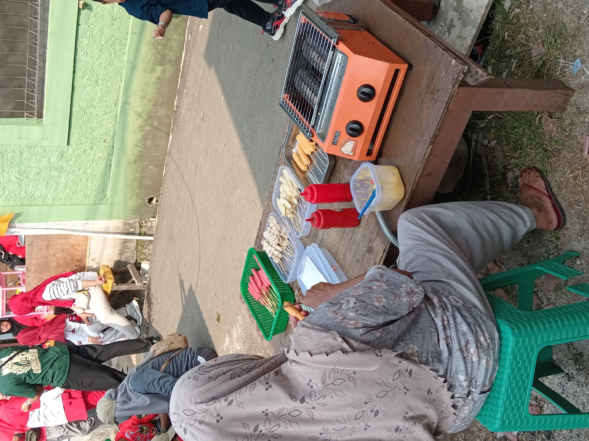 This image depicts an outdoor scene in Depok, Indonesia, likely a street or communal area, where a food vendor is preparing food. In the foreground, a woman wearing a grey hijab and a patterned top is seated on a green plastic stool, facing away from the camera. She is holding a skewer of food and appears to be preparing it.
On a rustic wooden table in front of her are various food items. A green basket contains numerous red sausages on skewers. Next to it, a clear plastic container holds lighter-colored, round food items on skewers, possibly meatballs. Another tray contains several longer, sausage-like items. Two red squeeze bottles, likely for condiments like ketchup or chili sauce, are also present. A small plastic container with a yellow substance and a blue utensil sits near the food.
Dominating the right side of the table is a portable orange gas grill. The grill is open, revealing hot charcoal inside and cooking grates. A woman's legs are visible on the far right, walking away from the table, suggesting other people are present in the vicinity.
In the background, other individuals are seen, some wearing red and white attire. They appear to be standing or sitting, some possibly attending an event or gathering. A textured green wall with a barred window forms the backdrop to the scene. The ground is paved, with some patches of grass and gravel visible. The overall impression is a casual, community-oriented setting, possibly during a local event or market day.