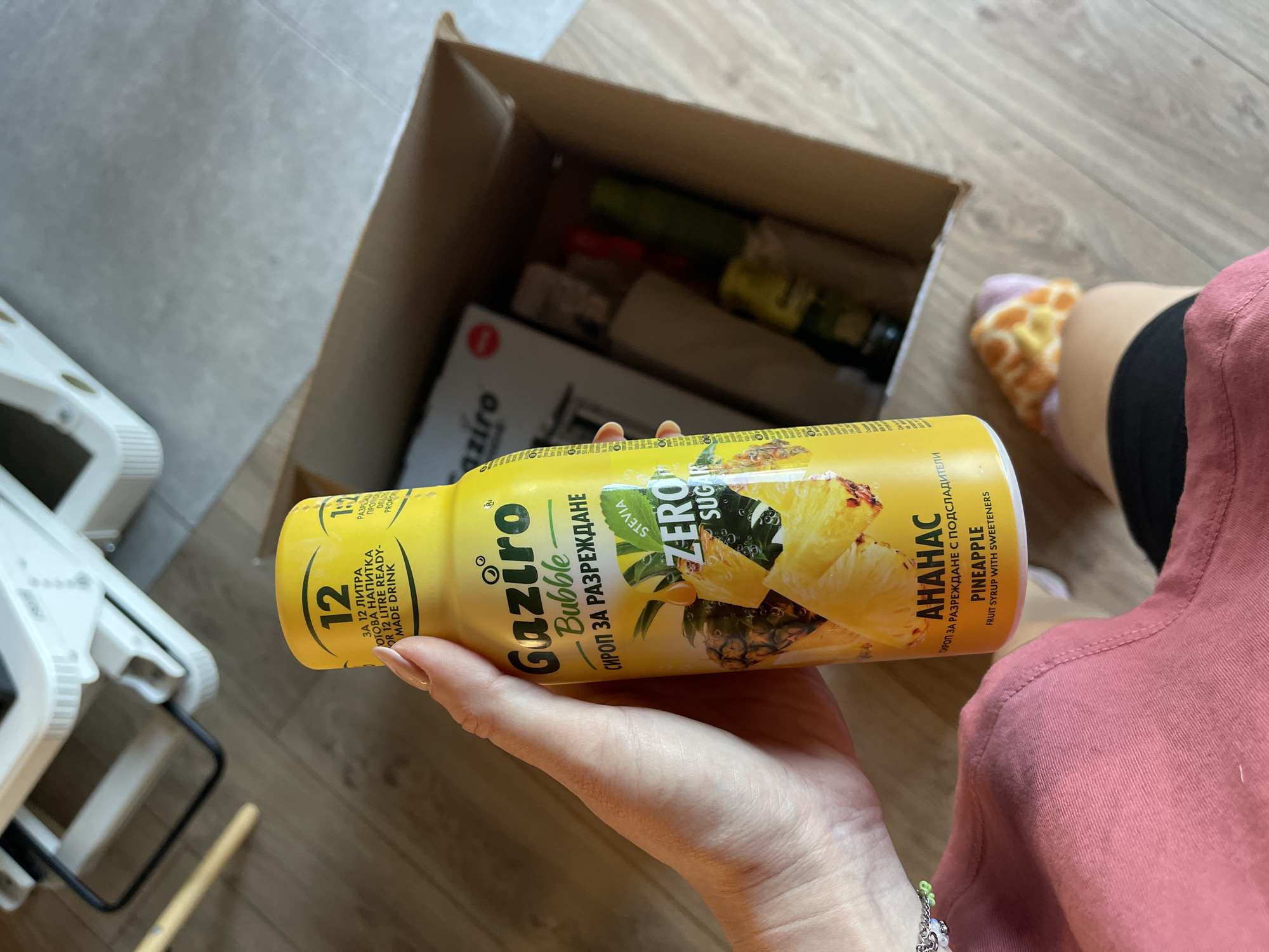 A person is holding a yellow bottle of "Gaziro Bubble" pineapple syrup in their right hand. The bottle is labeled "СИРОП ЗА РАЗРЕЖДАНЕ", which means "syrup for dilution" in Bulgarian. The bottle also states "ZERO SUGAR" and contains images of pineapples.
In the background, there is a cardboard box filled with various other bottles and items, suggesting that it might be a delivery box or a box of groceries. Also, a portion of a leg wearing a colorful slipper is visible.
The location is likely a private residence in Sofia, Bulgaria, given the branding of the product, and the items visible are common household goods. The presence of a baby chair to the left might indicate the presence of small children in the household.