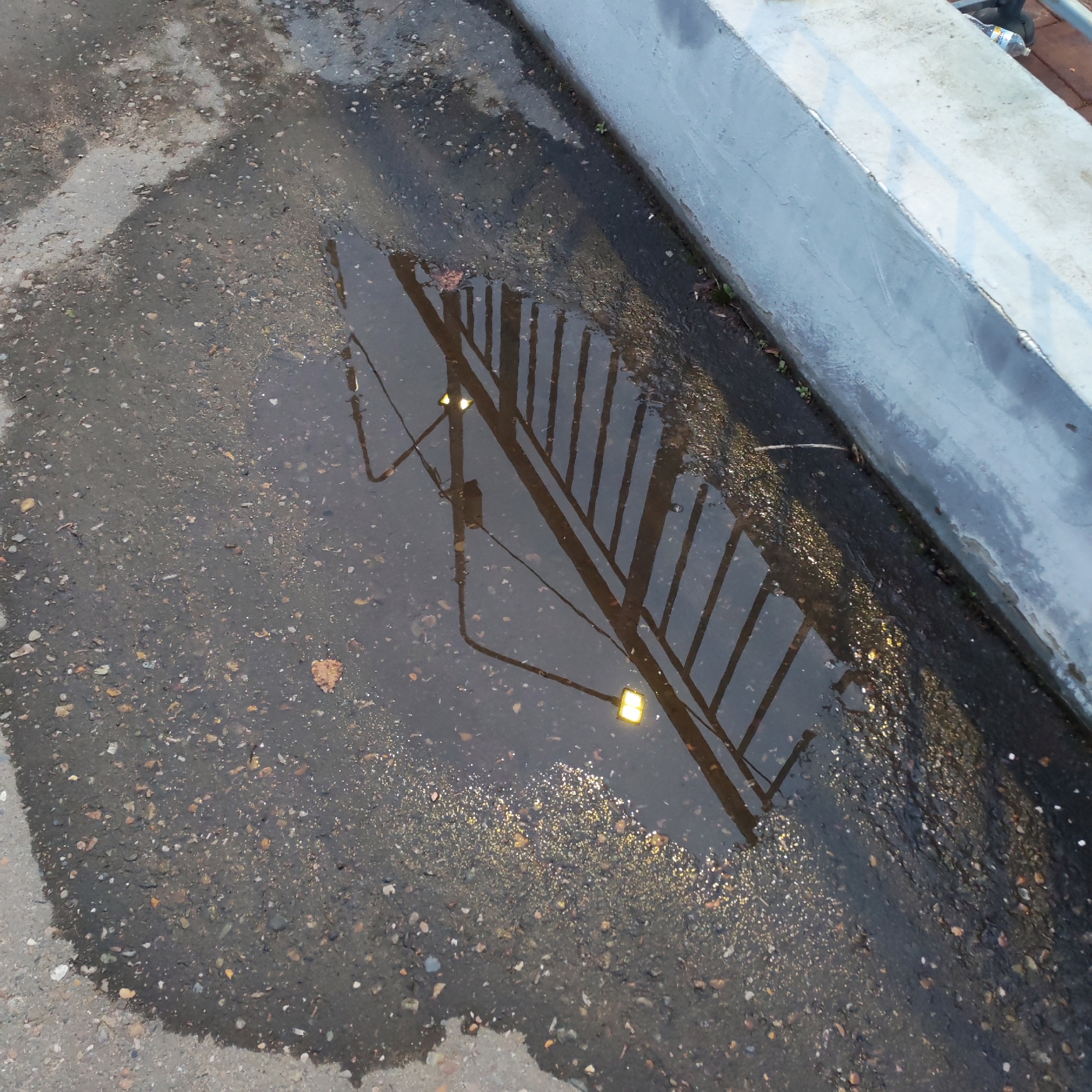A puddle on a paved surface reflects a railing and a street light. The puddle sits next to a concrete curb. The pavement has small rocks and pebbles embedded in it. The light reflecting in the puddle indicates a source of illumination. The photograph appears to have been taken in Chaikovsky, Russia.