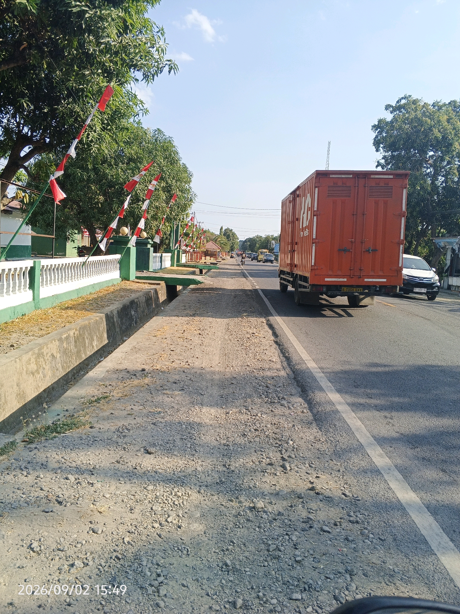 The image shows a street scene in Pinrang, Indonesia. A large orange truck is traveling on the right side of the road. On the left side of the road, there is a sidewalk with a concrete drainage ditch and a white fence with green trim. Red and white flags are strung along the fence and on trees. The sky is clear and blue. The timestamp on the image indicates the photo was taken on September 2, 2026, at 3:49 PM.
