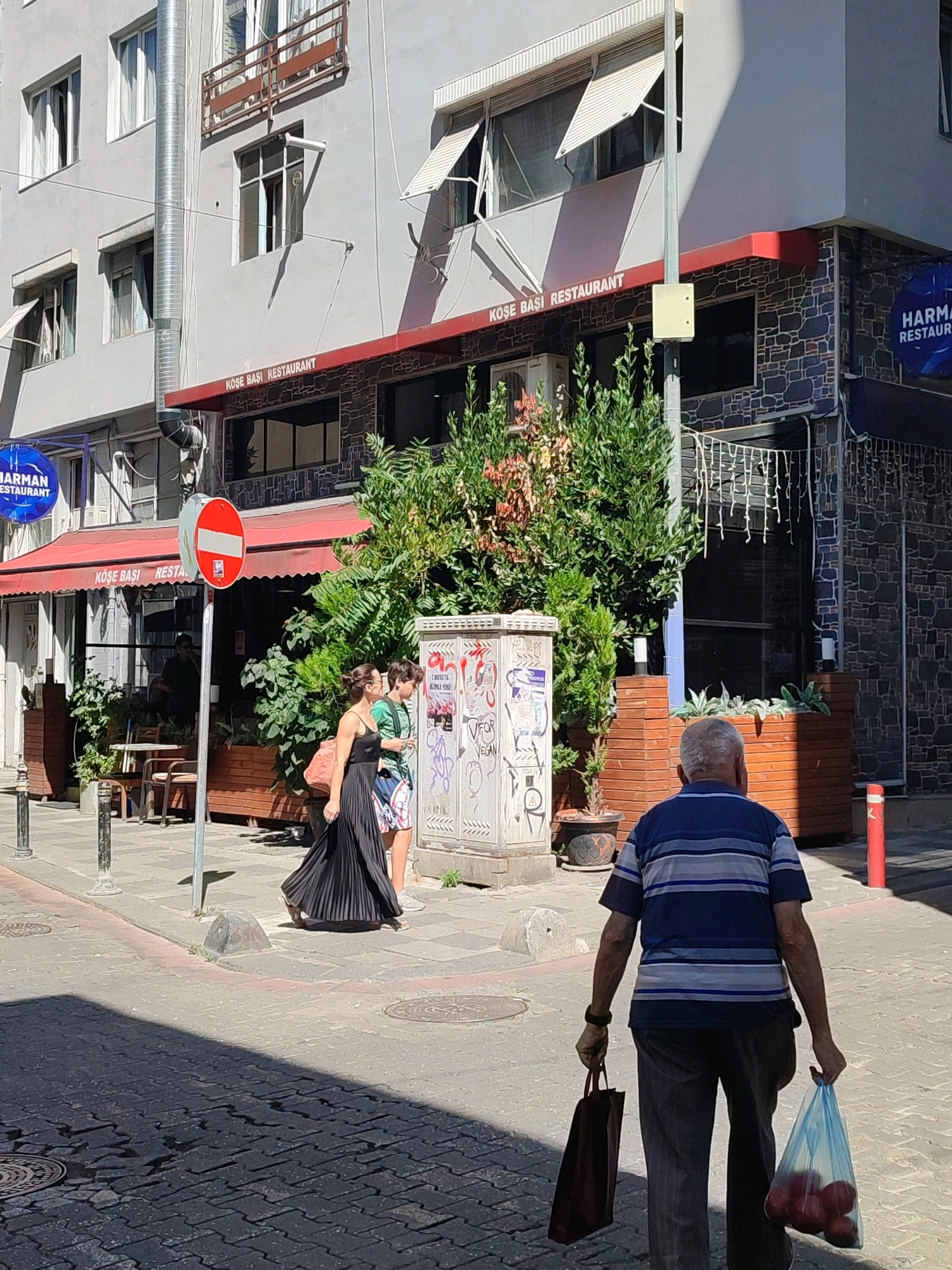 In Kadıköy, Turkey, on a sunny day, an elderly man with grey hair walks away from the camera, carrying a brown bag in his left hand and a transparent plastic bag filled with red apples in his right. He is wearing a blue and white horizontally striped t-shirt and dark trousers. Behind him, a woman in a long black dress and a younger person in a green t-shirt and patterned shorts walk away from the camera, appearing to be conversing. They are walking on a paved sidewalk in front of a building housing "KOŞE BAŞI RESTAURANT". The building has a grey facade with several windows and awnings. A red awning extends over the restaurant entrance. A blue circular sign with white text that reads "HARMAN RESTAURANT" is visible on the building. A red and white circular "no entry" sign is mounted on a pole in the foreground. The street is paved with dark cobblestones. Lush green plants are visible in planters and along the building's facade.