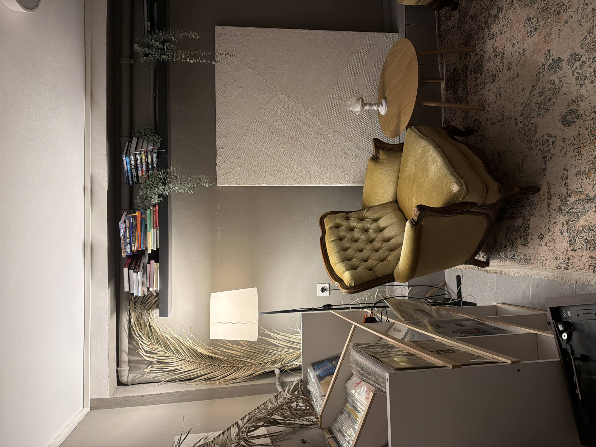 The image depicts an interior space with a vintage aesthetic. Dominating the scene is a tufted yellow-green armchair, situated beside a small round wooden side table. A textured abstract artwork hangs on the wall above. A built-in bookshelf filled with books is visible on the left side of the frame, with dried foliage adding a decorative touch. A floor lamp illuminates the corner, adjacent to a white shelf unit storing vinyl records. A patterned rug covers the floor. The location, Budva, Montenegro, cannot be verified from the visual information provided. No people or direct interactions are visible in the frame.