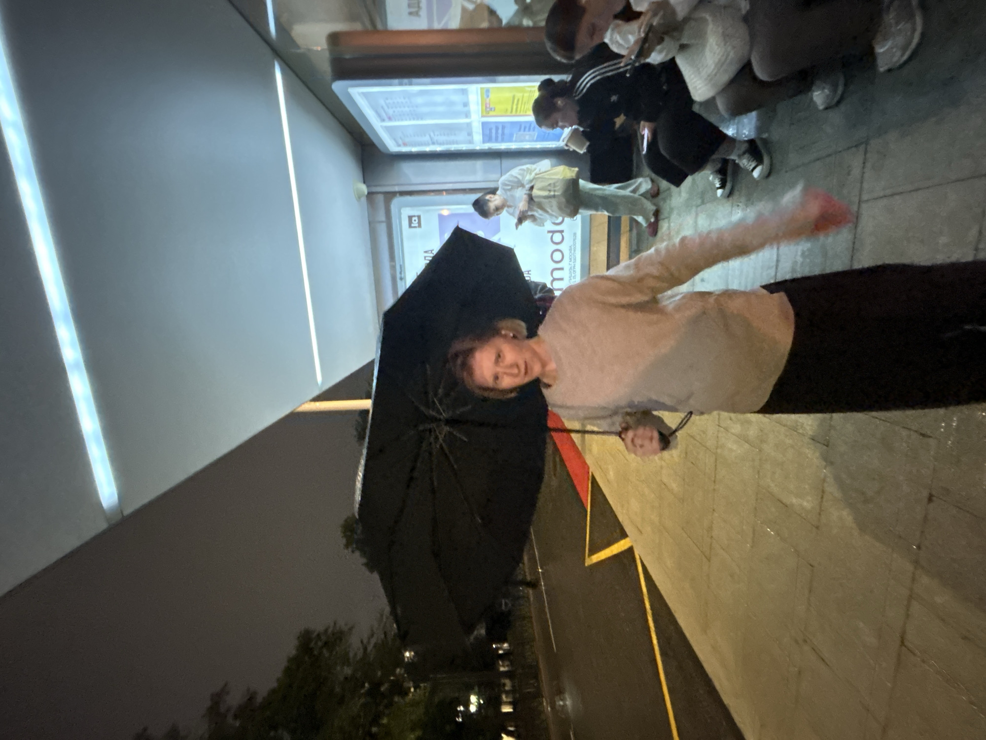 A woman stands under a large black umbrella at a bus stop at night. She is wearing a light-colored sweater and dark pants. Her umbrella is open, obscuring much of her body from the waist down. In the background, several other people are seated on a bench, some looking at their phones. A bus schedule is visible on a display board behind them. The ground is wet, indicating it has been raining. The bus stop has a modern, covered design with lighting. Trees and streetlights are visible beyond the bus stop.