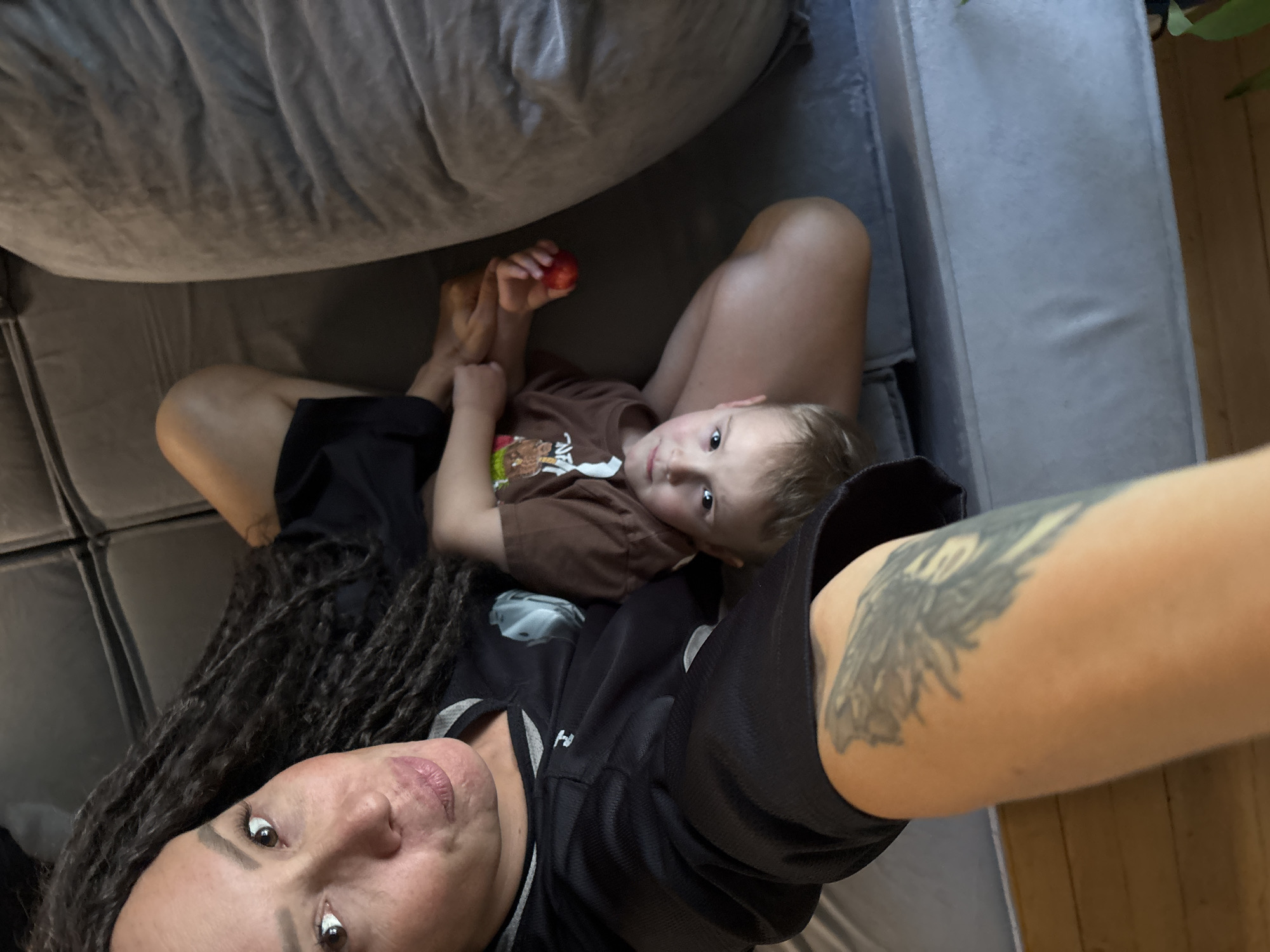 A high-angle selfie captures a woman and a young child relaxing indoors in Chaikovsky, Russia. The woman is in the foreground, lying on her back on a grey sofa, holding the camera to take the photo. She has long, dark dreadlocks and a large, dark, possibly tribal-style tattoo on her right arm. She wears a black and white athletic-style shirt. Her expression is neutral with a slight hint of a smile.
A young child with light hair is lying on her lap/chest, looking up at the camera with a neutral expression. The child is wearing a brown t-shirt with a colorful graphic, possibly depicting a bear, and holds a small red fruit, resembling an apple or plum, in their hands. The background shows the texture of the grey sofa cushions, suggesting a comfortable, domestic setting. A glimpse of a wooden floor and a green plant is visible on the far right. The overall atmosphere is relaxed and candid, indicating a moment of quiet time together. No discernible text is visible on clothing or in the background.