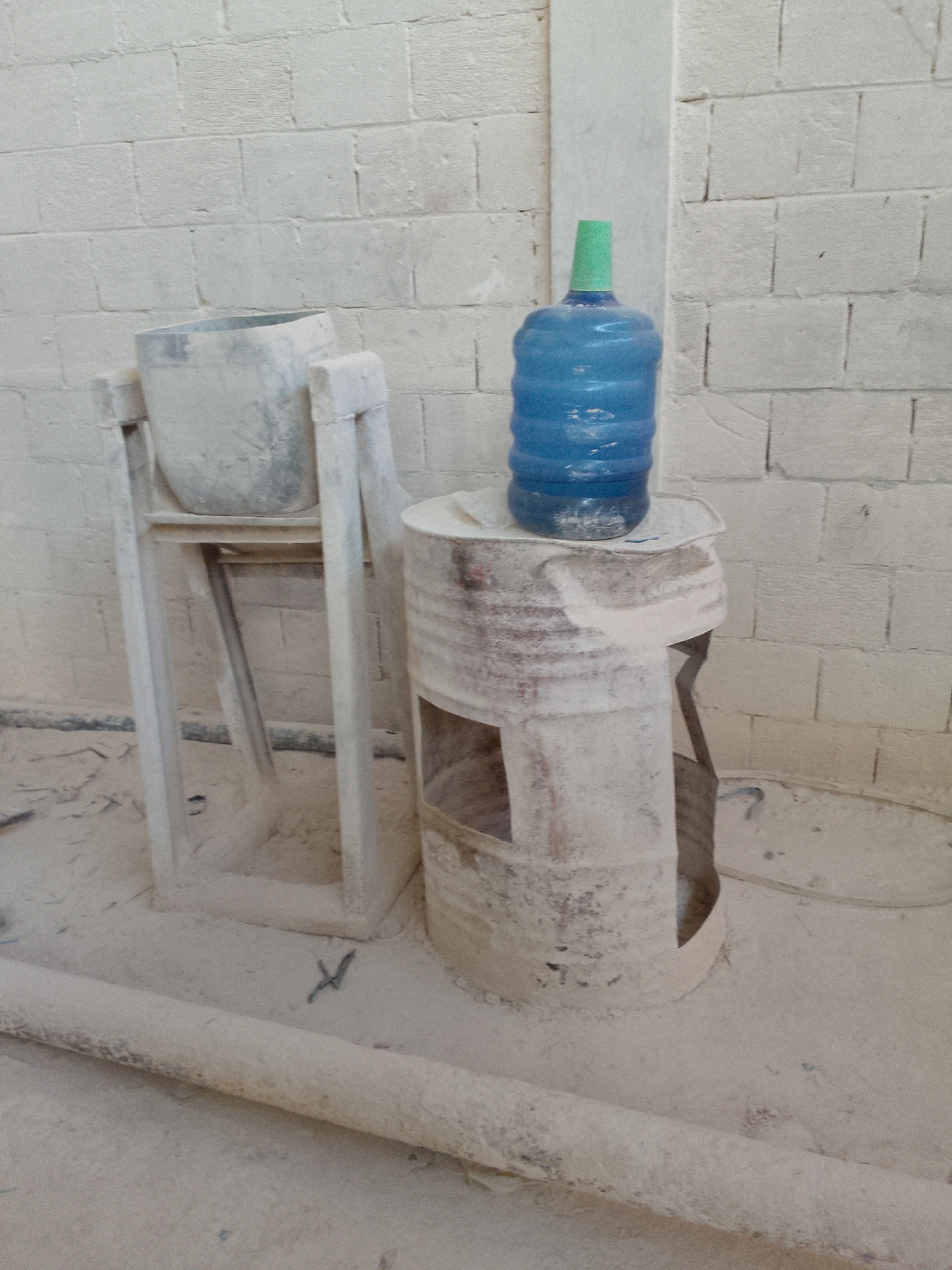 In Gunungmasigit, Indonesia, a scene unfolds within a dusty, white-bricked room. In the foreground, a thick, horizontal pipe, also covered in dust, lies on the floor. To the right, a repurposed metal drum, with cut-out sections revealing an inner cavity, serves as a stand for a large blue water jug with a green cap. The jug is partially filled with liquid. To the left, a metal basin sits atop a simple, two-legged stand, both also dusted. The overall impression is one of a utilitarian or construction setting, with the pervasive dust suggesting recent or ongoing work.