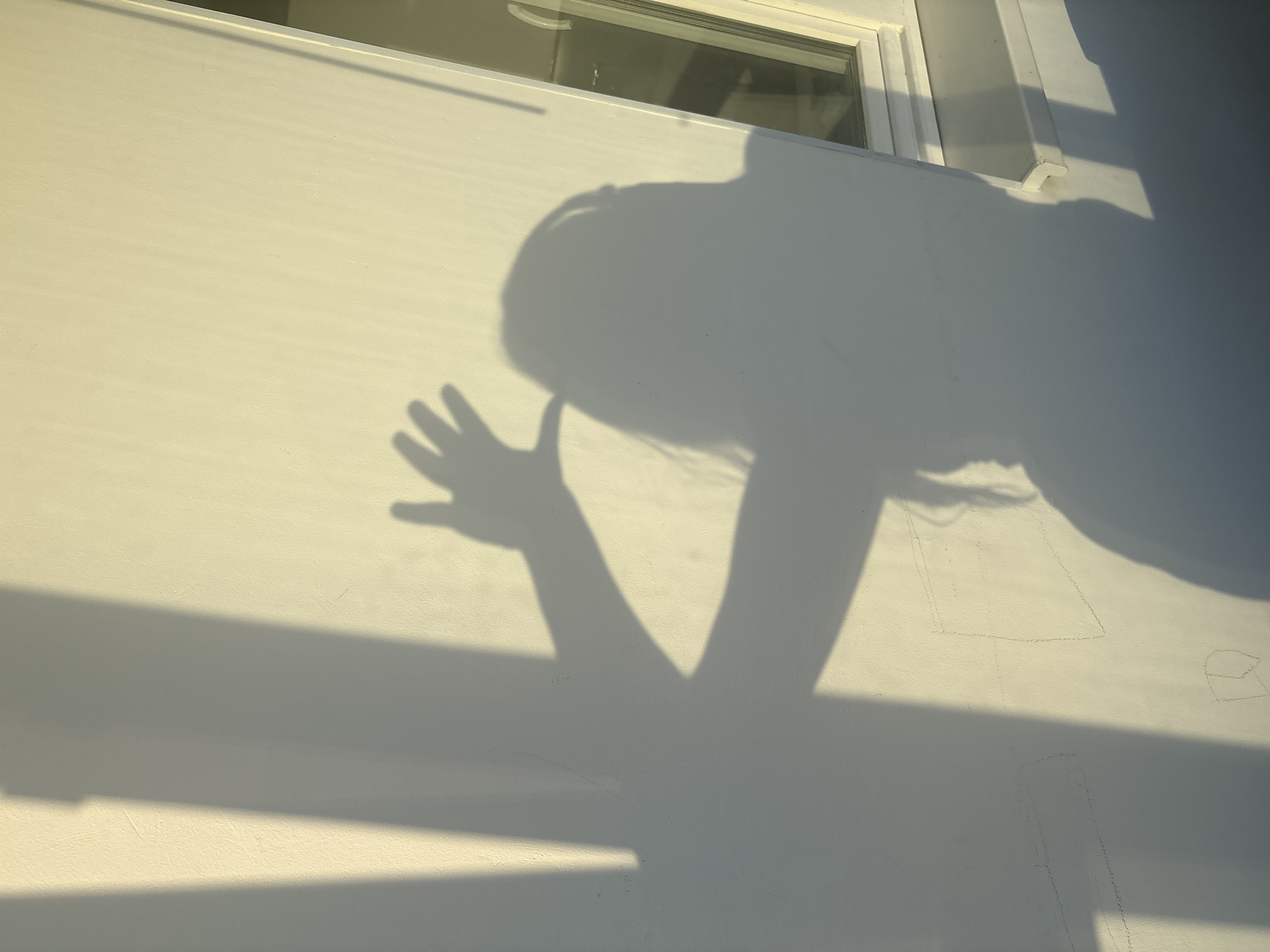 The image shows a shadow of a person with long hair and an outstretched hand casting onto a light-colored wall. The shadow is positioned in the center-right of the frame, with a second, fainter shadow to the left, possibly from a different object or person. A portion of a window frame and sill is visible on the right side of the image. The lighting suggests it is either sunrise or sunset, casting long shadows. The scene is not discernible as being in Chaikovsky, Russia, as there are no identifying landmarks or contextual clues present.