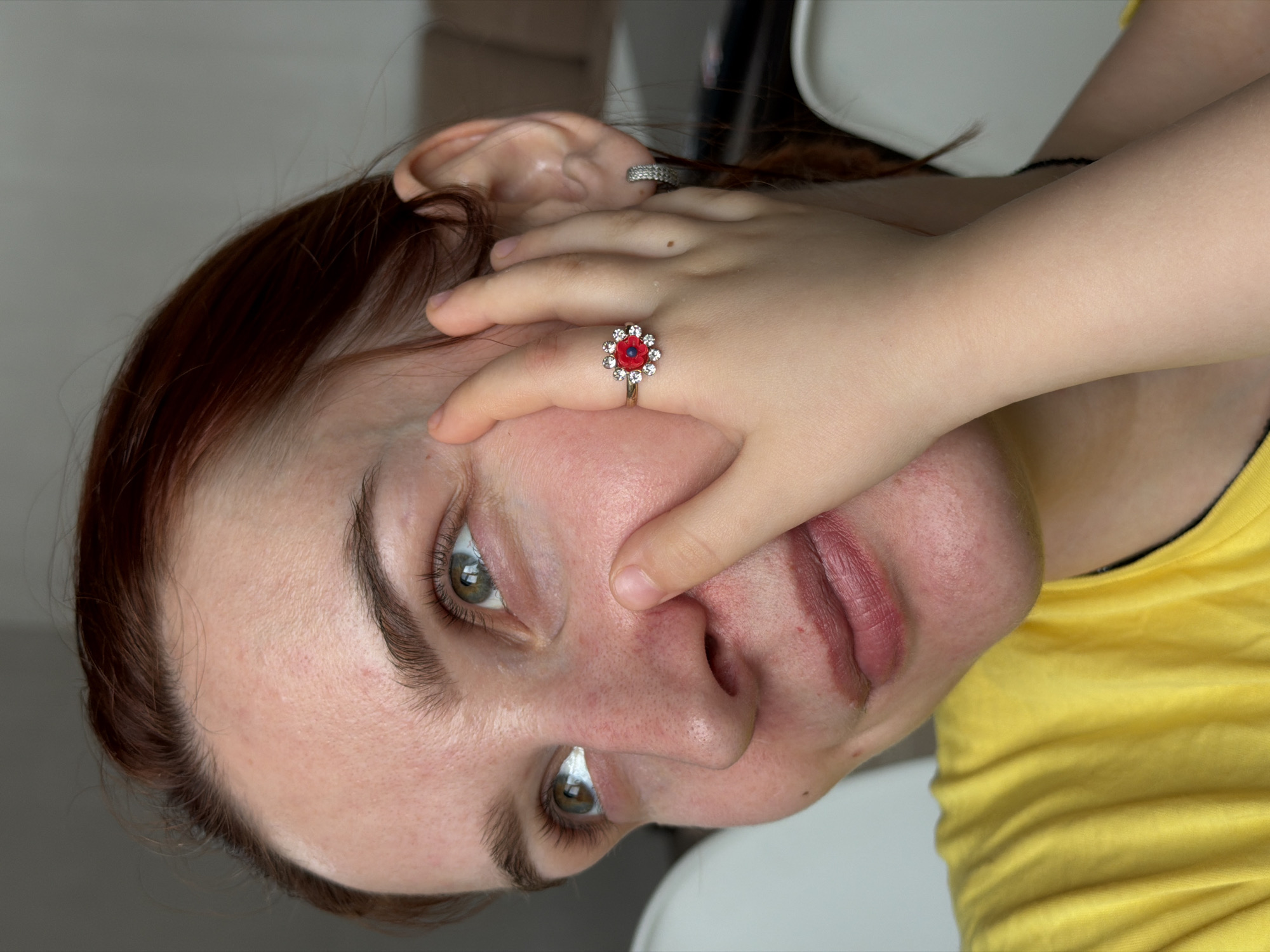 A close-up, rotated image features a woman with auburn hair wearing a yellow shirt. Her head is angled, revealing her face, neck, and the upper part of her torso. A child's hand, adorned with a red and white flower ring, partially covers the woman's face, obscuring her nose and part of her mouth. An earring is visible on her left ear. The background is a blurred mix of white and indistinct shapes. There is no indication in the image that it was taken in the city of Chaikovsky, Russia.