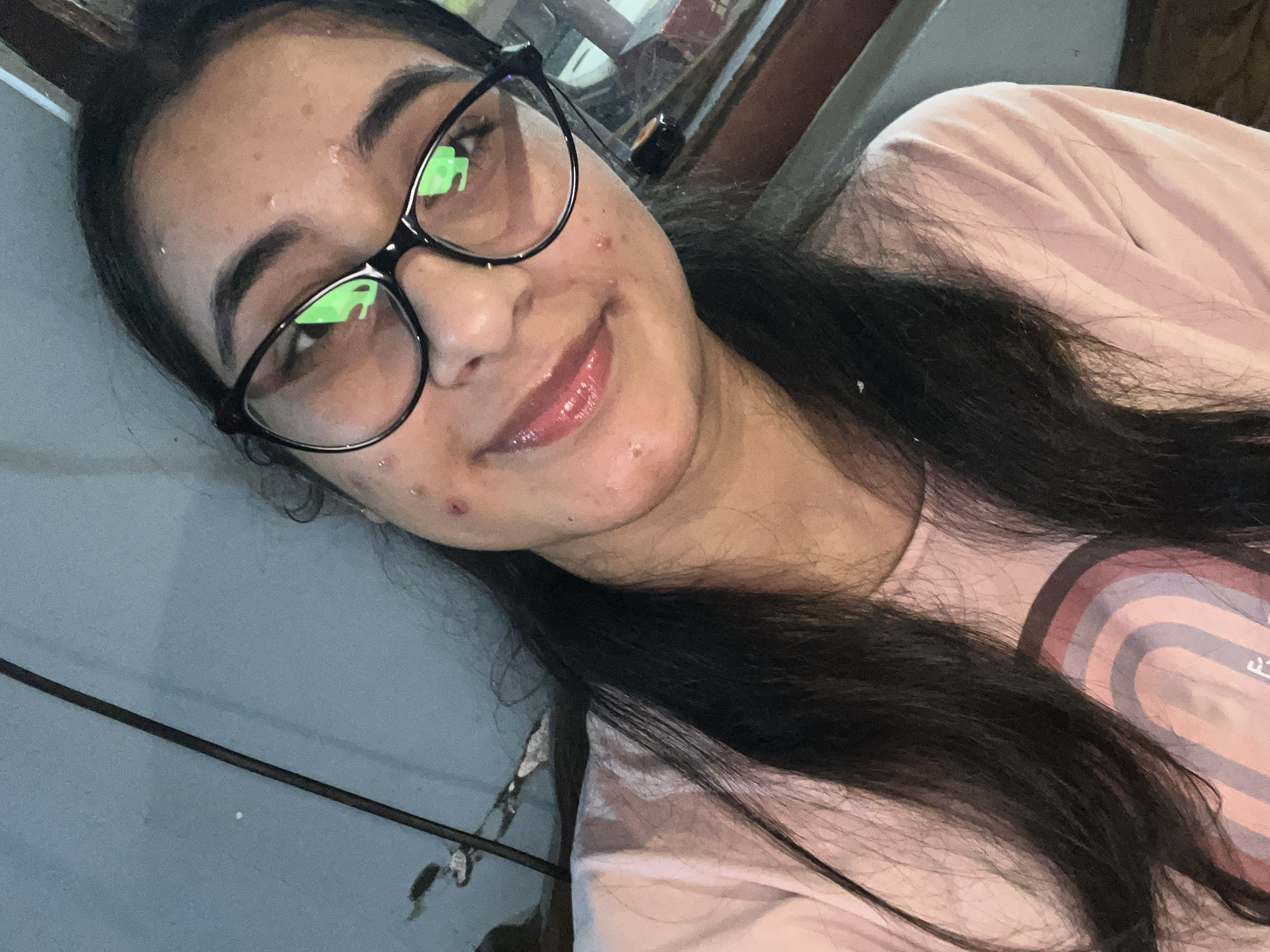 A young woman with long, dark hair is looking at the camera and smiling. She is wearing a pink t-shirt and black-rimmed glasses. Her skin has several blemishes, and she is wearing lip gloss. The background is a blue wall, with some cables and what appears to be furniture visible. The lighting is dim, suggesting the photo was taken indoors.