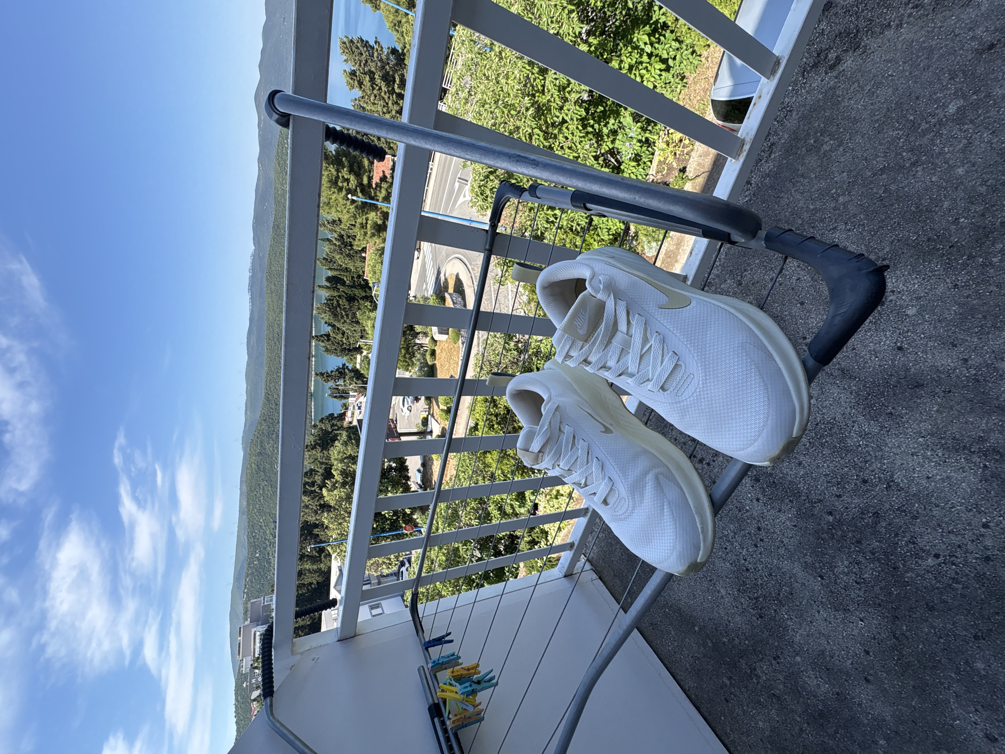 Two white Nike sneakers are drying on a metal clothes drying rack on a balcony. The background shows a view of Neum, Bosnia and Herzegovina, including water, trees, buildings, and a road. Several clothespins are attached to the lower portion of the drying rack.