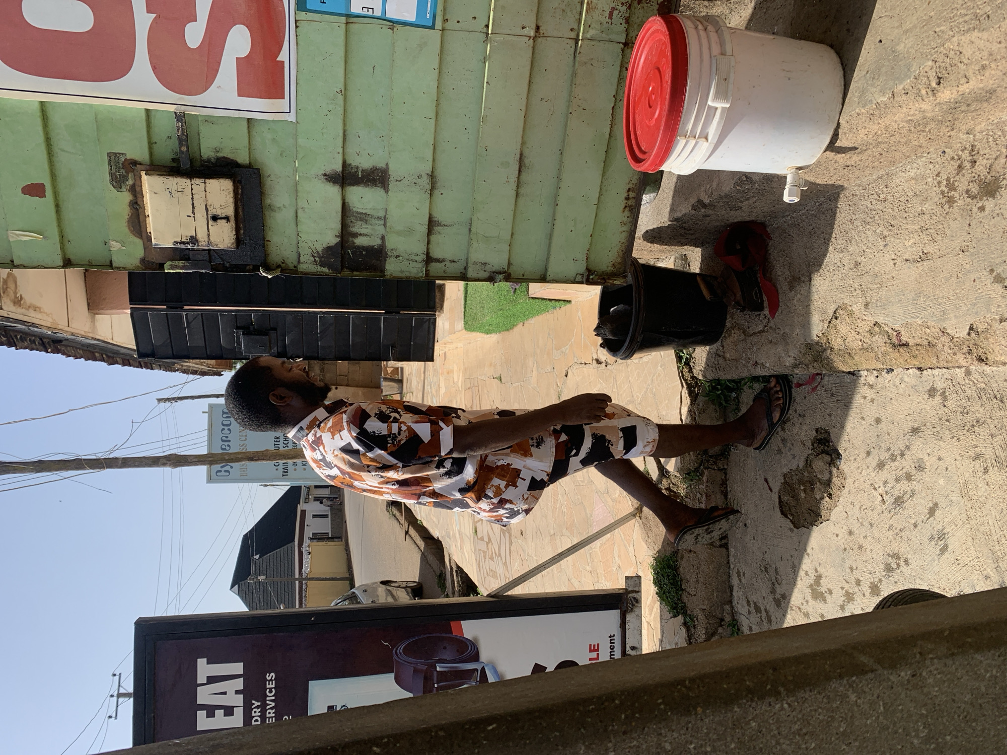 A man is standing on a sidewalk in Jos, Nigeria. He is wearing a patterned shirt and shorts and sandals. To his right, there is a green building with a metal door and a sign in red letters. There is a black bin and a white bucket with a red lid on the ground beside the building. To his left, there is a black sign with the word "EAT" on it, followed by "DRY SERVICES". Above that, there is a sign for "Cybercom Business Center". In the background, there are trees and buildings. The sidewalk is made of concrete and has some cracks and stains.