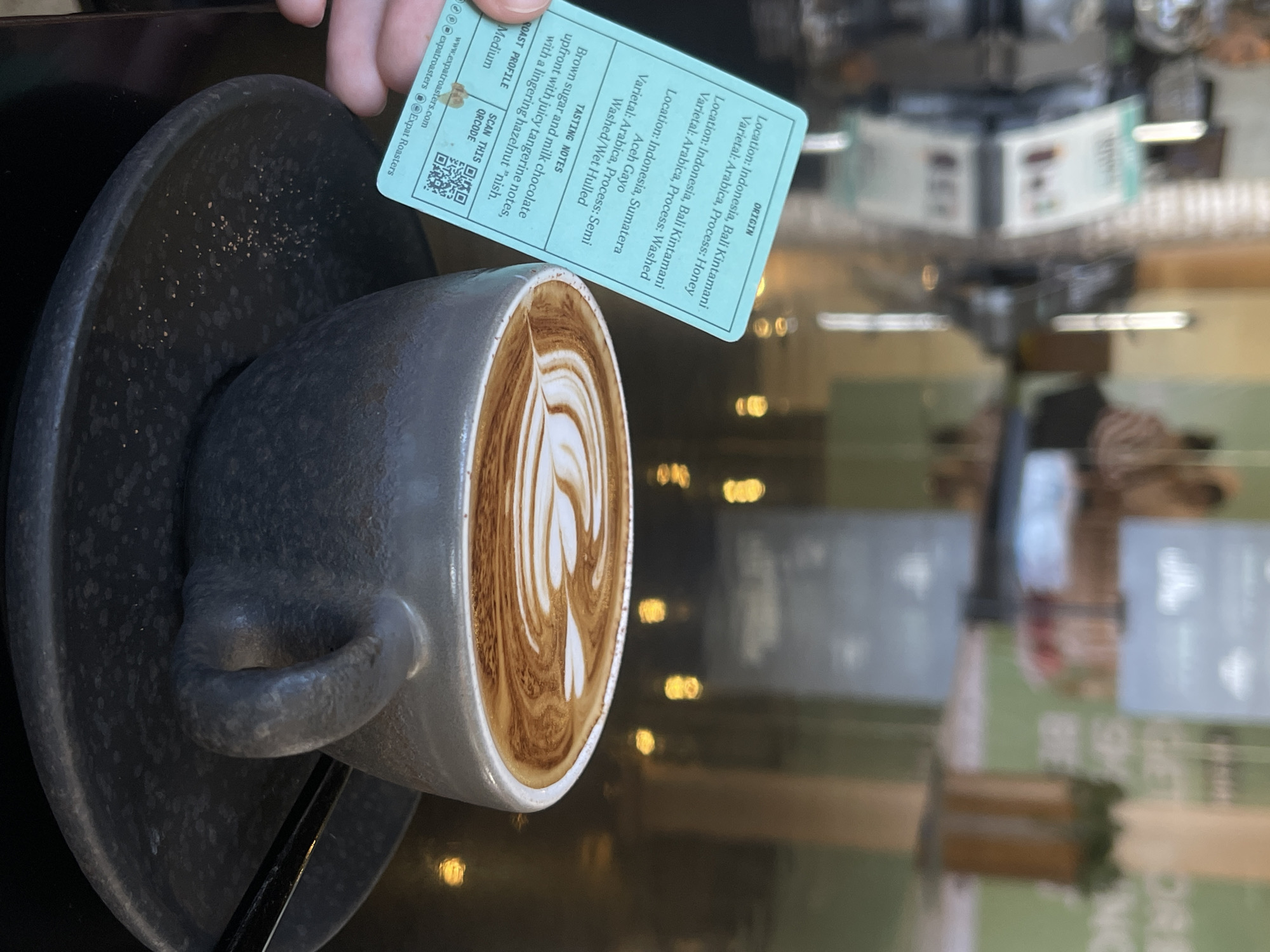 A close-up, vertical shot captures a cup of coffee with intricate latte art on a dark, textured saucer, placed on a highly reflective black table. A hand holds a light blue information card next to the cup, providing details about the coffee beans.
The card, from "Expat Roasters," indicates the origin of the coffee beans. It lists three primary origins: Indonesia, Bali Kintamani (with both Honey and Washed processes for Arabica varietal), and Indonesia, Sumatera Aceh Gayo (with a Semi Washed/Wet Hulled process for Arabica varietal). The tasting notes describe the coffee as having "Brown sugar and milk chocolate upfront with juicy tangerine notes, with a lingering hazelnut 'nish.'" The roast profile is "Medium." The card also displays a QR code to "SCAN THIS" and the website "www.expatroasters.com" along with "@expatroasters" for social media.
The coffee cup is a dark, possibly charcoal-grey, ceramic or stoneware mug with a sturdy handle, matching the saucer which has a subtle speckled texture. The latte art features a symmetrical, multi-layered heart or rosetta pattern, indicating a skilled barista. The surface of the table reflects blurred lights and shapes from the background, suggesting an indoor cafe setting in Kuta, Indonesia, as contextually provided. The reflections create a luminous, atmospheric effect, blurring any specific time of day but suggesting ambient lighting.
