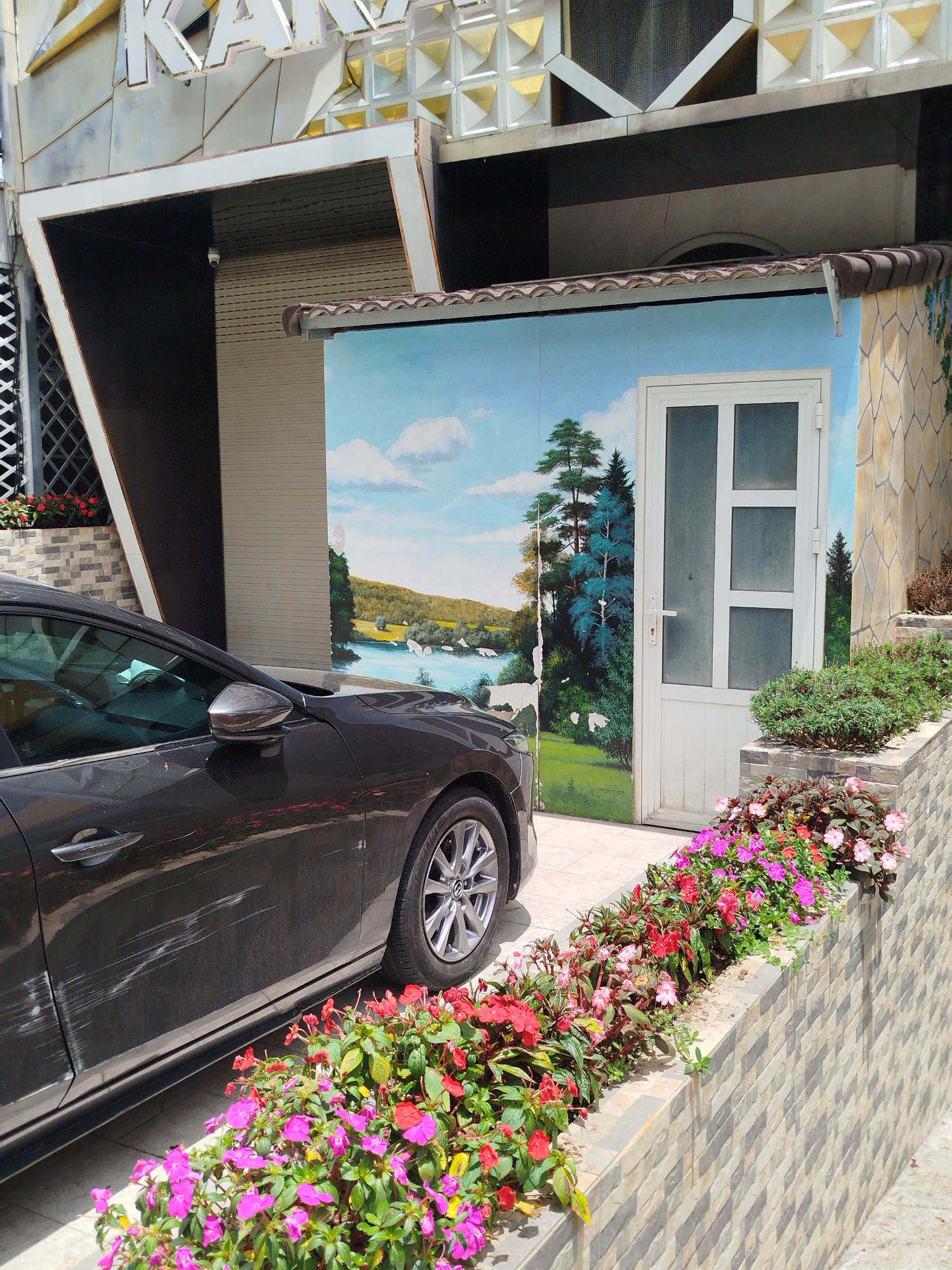 A dark-colored sedan is parked in front of a building with a mural painted on its side. The mural depicts a landscape with a body of water, trees, and hills under a cloudy sky. A white door with glass panels is visible within the mural. In the foreground, a wall is decorated with colorful flowers, including pink and red impatiens, and green foliage. The building above the mural has an angular, metallic facade with geometric patterns. A security camera is mounted on the building's exterior. The scene appears to be in an outdoor setting in a city, possibly Lâm Viên - Đà Lạt, Vietnam, given the architectural style and the presence of flowers commonly seen in the region. The car's side shows visible scratches and scuff marks.