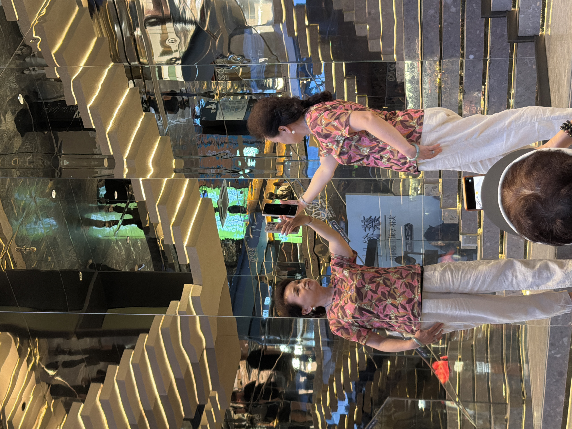 In Yuzhong District, China, a middle-aged East Asian woman is captured in a reflective setting, possibly a modern architectural space with extensive mirrored surfaces and illuminated tiered structures. The woman stands in the foreground, holding up a smartphone with her right arm extended, seemingly taking a selfie or recording a video. She is wearing a short-sleeved, collared shirt with a vibrant pink and floral pattern, paired with loose, light-colored linen pants. Her left hand rests on her hip. A second woman, also East Asian and middle-aged, is visible in the reflection on the left, mirroring the first woman's pose and attire, holding a similar smartphone. The environment features a dynamic interplay of light and reflection, with stylized, illuminated stairs or shelves forming geometric patterns that repeat across the mirrored walls and ceiling. The overall impression is one of a visually complex and contemporary urban environment.