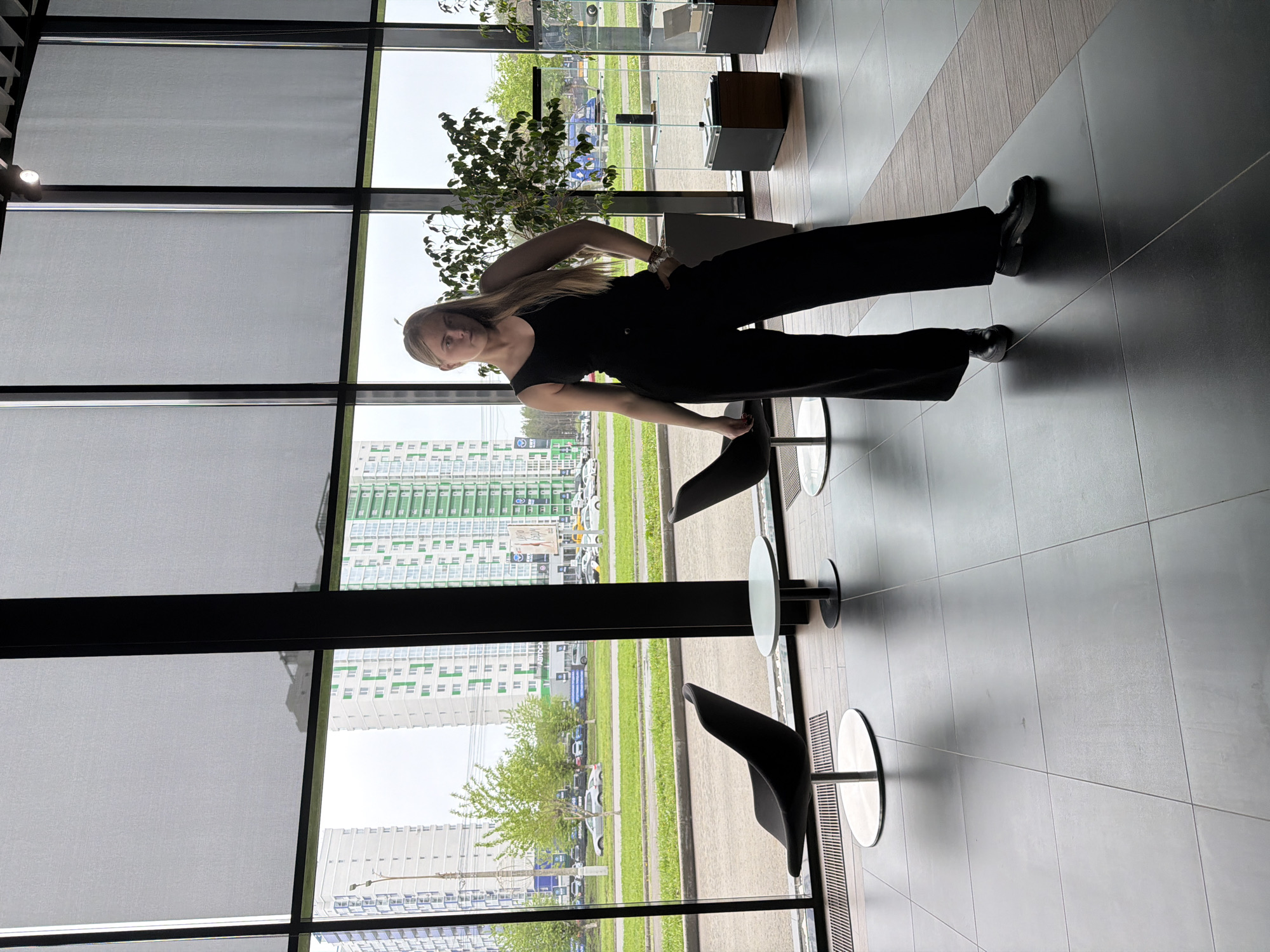 A young woman is standing inside a building with a large window overlooking a cityscape. She is wearing black attire, including a sleeveless top and wide-leg trousers. Her blonde hair is partially tied back.
Outside the window is an urban view, with modern high-rise apartment buildings in the background, and cars parked on the street. A small tree is also visible in the foreground of the outdoor scene. The window has horizontal blinds.
Inside the building, there are tables with round tops and black chairs placed next to the window. The floor is tiled with dark gray tiles. The building appears to be located in Izhevsk, Russia.