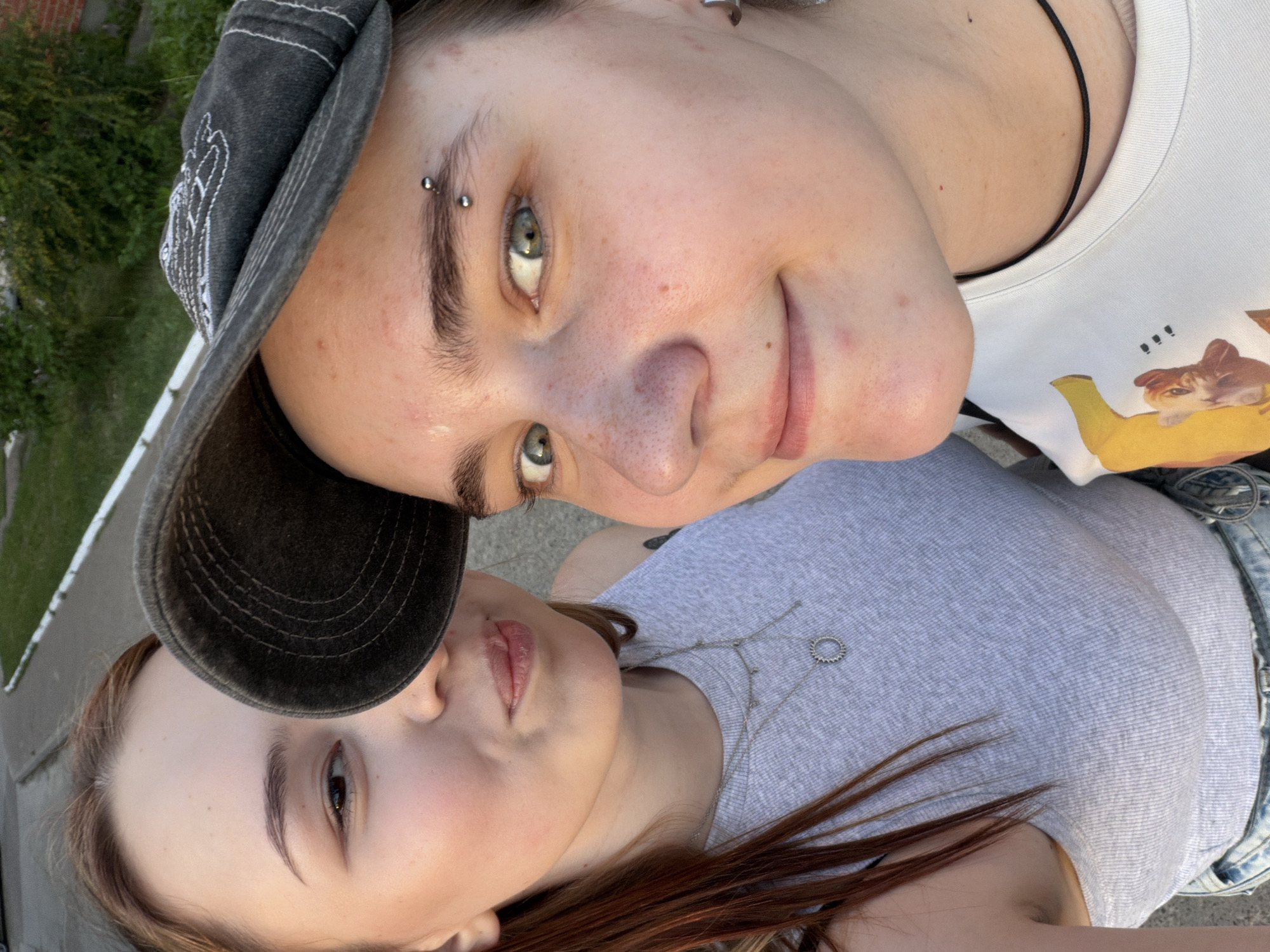 Two people are close to each other, with one person wearing a baseball cap and the other without. The person in the cap has an eyebrow piercing and is wearing a white t-shirt with a graphic of a cat. The other person is wearing a grey shirt and a necklace. The location appears to be outdoors. There is no other information.
