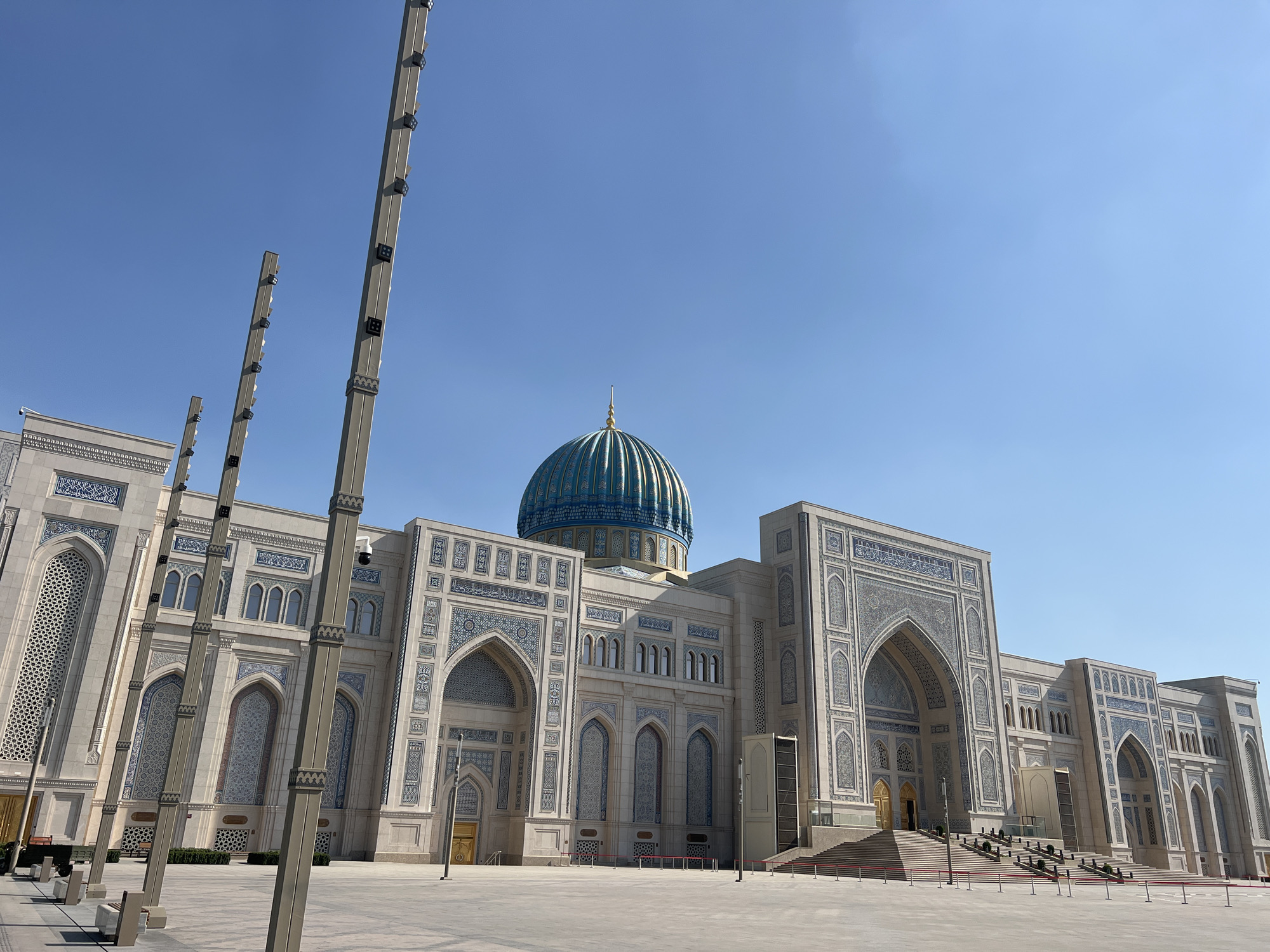 The image depicts the exterior of a grand building, likely a mosque given its architectural style, under a clear, bright blue sky. The building's facade is intricately decorated with blue and white geometric patterns and Arabic calligraphy. A prominent turquoise dome with gold accents rises from the center of the structure. Tall, slender poles with lights are positioned in the foreground, leading the viewer's eye towards the building. The ground in front of the building is a large, paved plaza. Red carpeting lines the steps leading up to the main entrance on the right side of the building. The scene appears to be set during daytime, with ample sunlight casting shadows, suggesting good weather. There are no people visible in the image, giving a sense of quiet grandeur. The architectural style, particularly the use of intricate tilework and the dome, is characteristic of Central Asian Islamic architecture. The location cues, combined with the architectural style, point to Tashkent, Uzbekistan. The overall impression is one of majestic scale and intricate craftsmanship.
