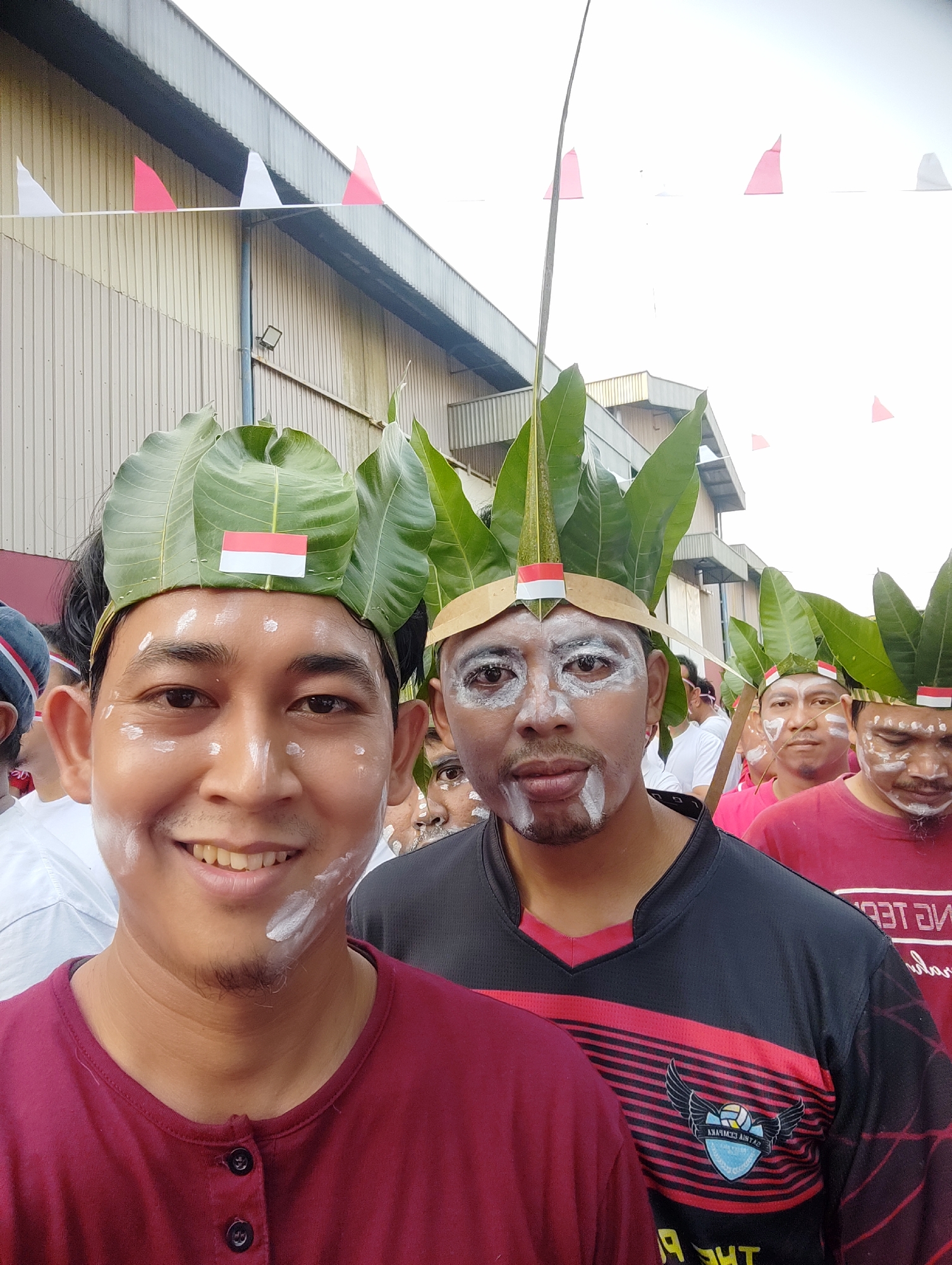 In a selfie-style shot, a group of men, likely participating in a cultural or nationalistic event, are gathered outdoors. The foreground is dominated by two men wearing elaborate headpieces made of large green leaves, resembling crowns. They have white paint applied to their faces, with some men having more extensive markings around their eyes and mouths. A small Indonesian flag is affixed to the headpiece of the man on the left. The man on the left wears a maroon henley shirt, while the man next to him wears a black and red athletic shirt with a logo. In the background, other individuals are visible, also adorned with similar leaf headpieces and face paint. Triangular flags in red and white, reminiscent of Indonesian national colors, are strung along a building in the upper portion of the image. The building itself appears to be a large, industrial structure with a corrugated metal exterior and a long, sloped roof. The overall atmosphere suggests a festive or commemorative occasion taking place in an outdoor urban setting, possibly within an industrial area of East Jakarta, Indonesia, given the architectural style of the building.