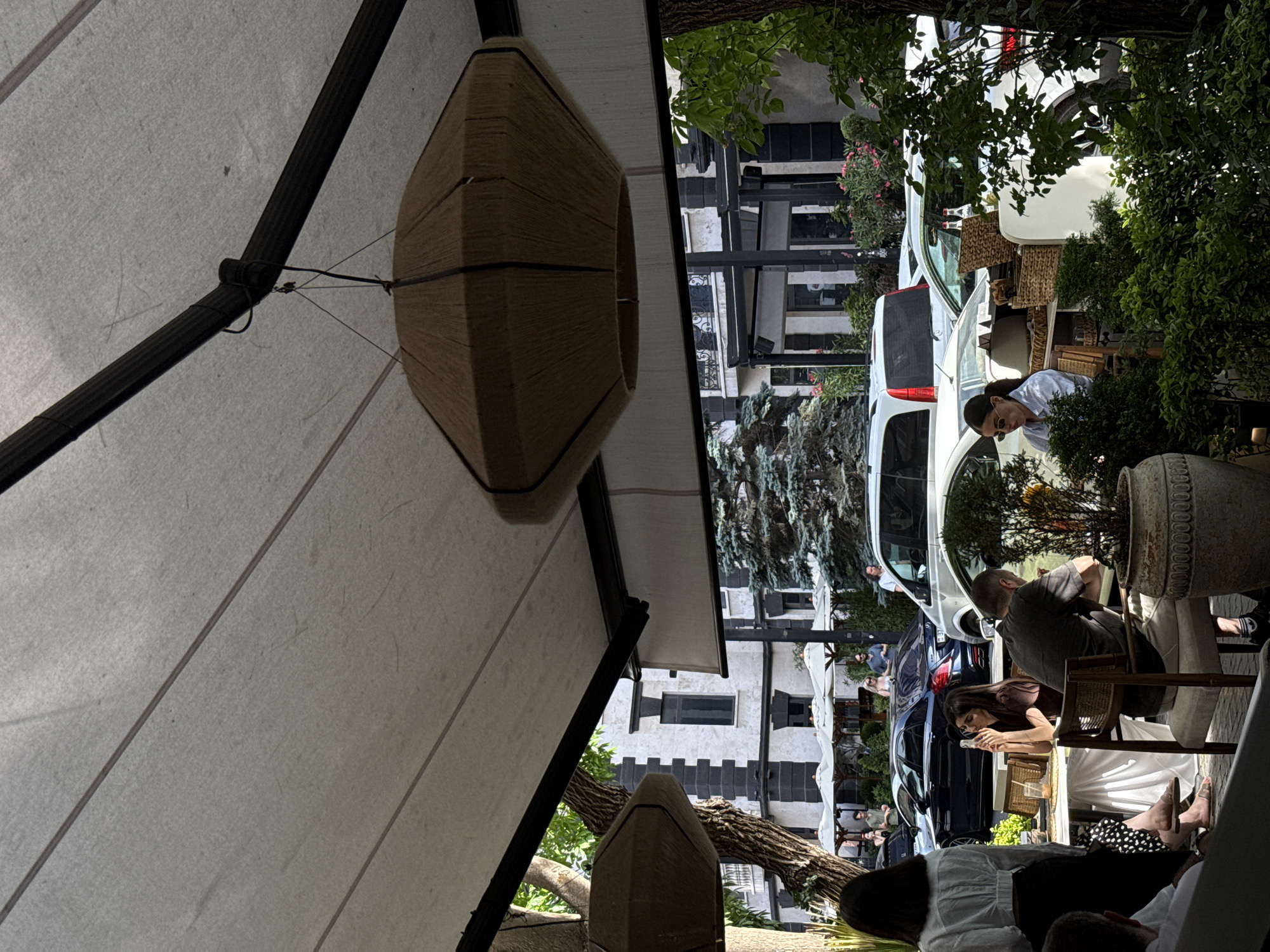 The image depicts an outdoor seating area, likely a cafe or restaurant, on a sunny day in Yerevan, Armenia. The scene is characterized by white canopies providing shade, a prominent woven pendant light fixture, and lush greenery. Several people are visible, some seated at tables and others near parked cars.
In the foreground, a woman with dark hair and sunglasses is seated, looking down at her phone. Further back, a man wearing a gray t-shirt is seated at a table, and another woman with long dark hair is also seated, engrossed in her phone. A dark sedan and a white SUV are parked on the street behind the seating area, suggesting an urban setting. The architecture in the background features white buildings with dark trim and windows, consistent with urban architecture. The presence of trees and decorative plants adds to the outdoor ambiance.
The lighting suggests it is daytime, with bright sunlight casting shadows. The overall mood appears relaxed, with individuals engaged in personal activities or enjoying the outdoor atmosphere. No visible text is present in the image.