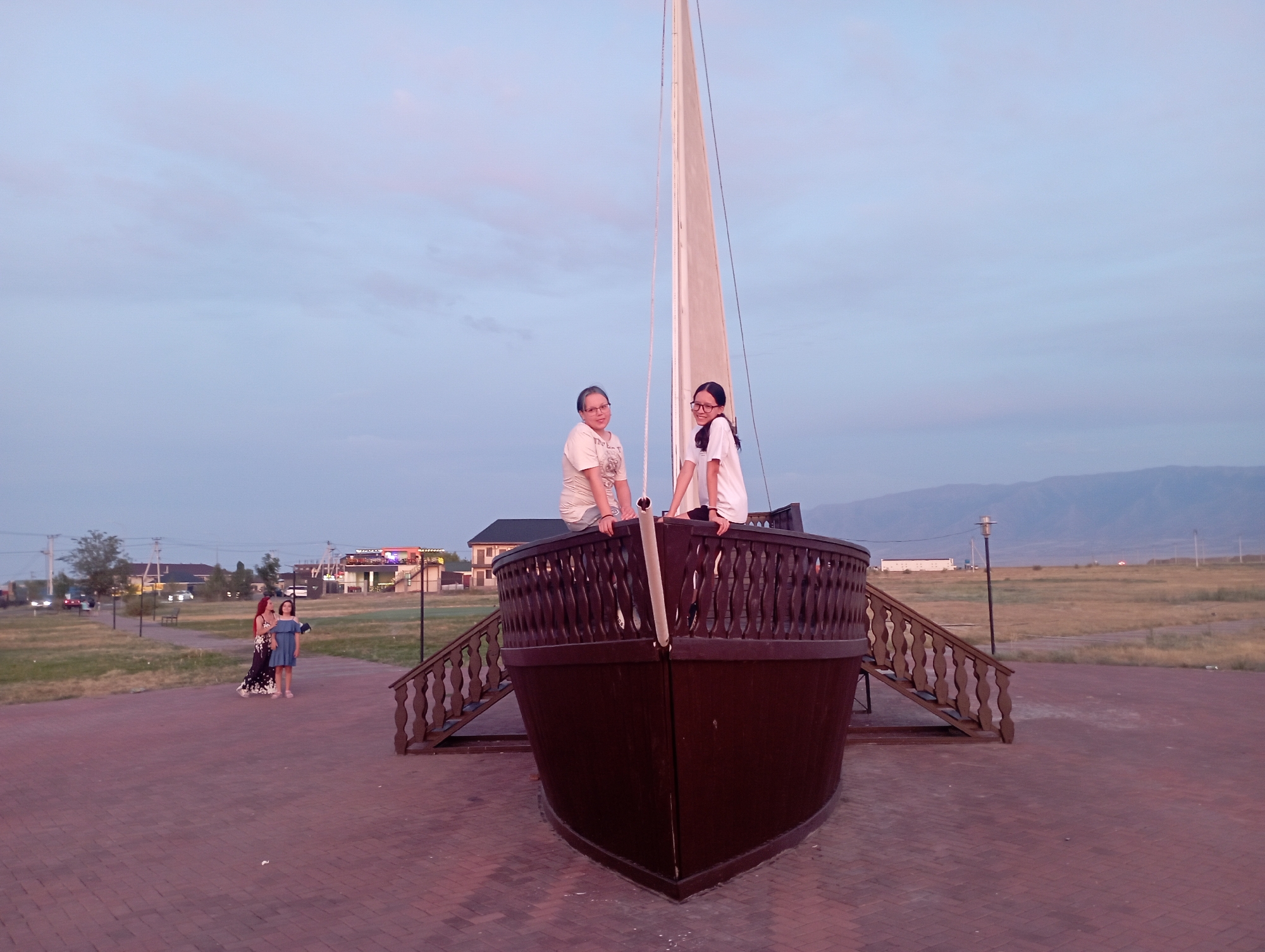 In the city of Unknown, Unknown, two individuals, an adult and a child, are seated on the railing of a wooden ship replica. The adult, wearing a light-colored t-shirt and glasses, is on the left, and the child, in a white t-shirt and glasses, is on the right. A tall mast rises behind them, with a sail furled. Two other individuals, a woman in a dark floral dress and a young girl in a blue dress, are standing on a brick pathway to the left of the ship. The ship replica is positioned on a paved area in front of a grassy field. In the background, there are buildings, streetlights, and a range of distant mountains under a pale blue sky with wispy clouds. The overall lighting suggests it is dusk.