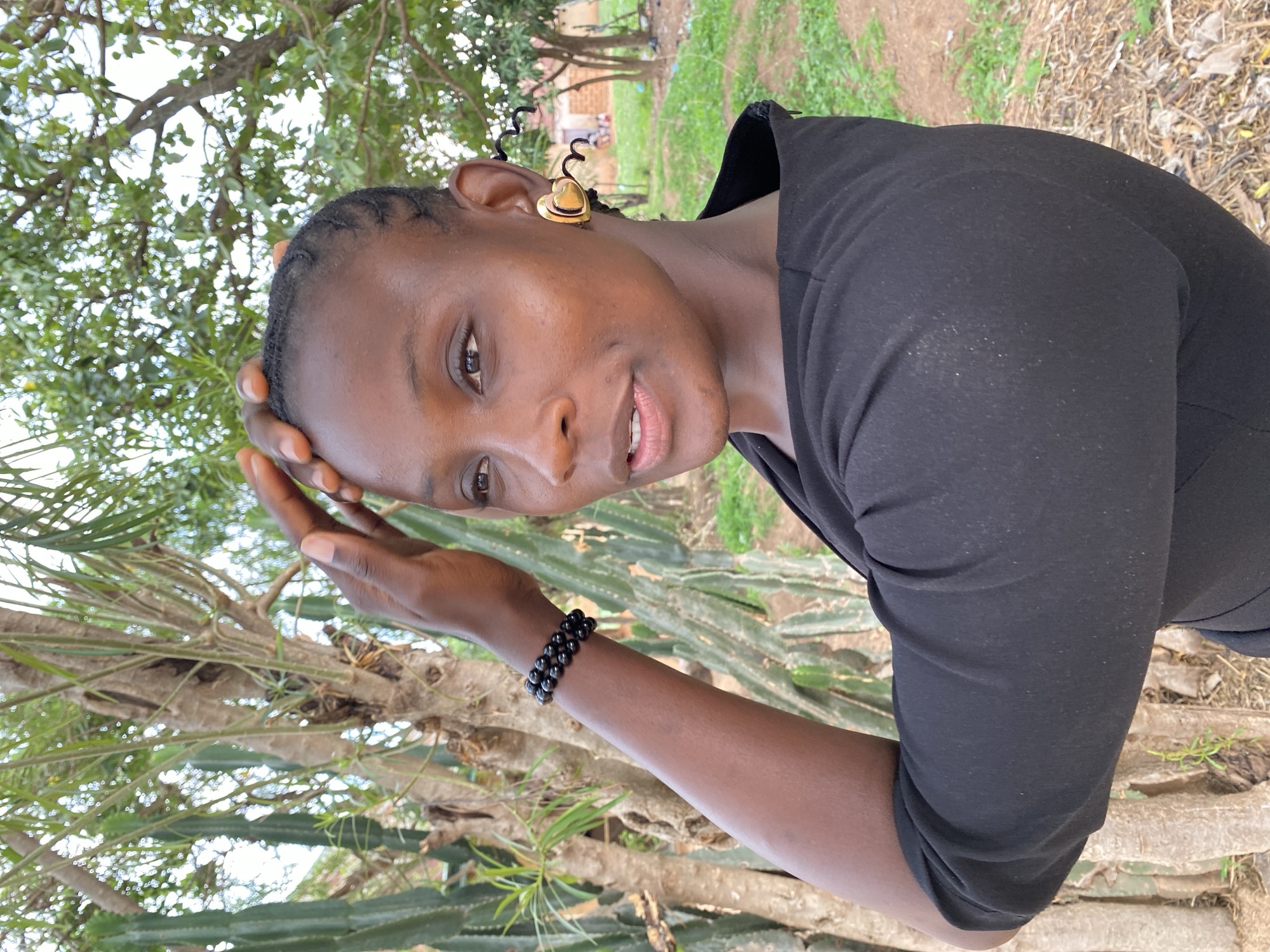 A dark-skinned woman with short braided hair and a gold heart-shaped earring with a curly black wire extension poses outside near a cactus-like tree. She wears a black fitted shirt, and a black beaded bracelet adorns her wrist. She touches her forehead with her right hand. The location appears to be in Kuru Dungus, Nigeria, as indicated by the prompt. The background shows greenery, including trees and grass. A glimpse of a building is visible in the far background. The ground consists of grass and dry leaves.