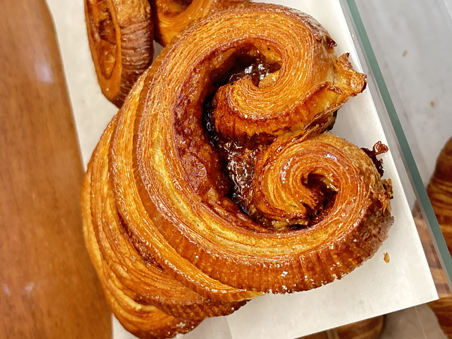 A close-up, high-angle photograph presents a single, large, golden-brown laminated pastry, consistent with a caramelized Kouign-amann, displayed within a bakery or cafe in Tel-Aviv, Israel. The pastry, roughly oval in shape, exhibits multiple distinct, glossy layers of dough spiraling inwards, forming two prominent central swirls that contain a dark, sticky, caramelized filling. Its surface is deeply caramelized to an amber-brown hue, featuring a shiny, sugary crust with visible granular sugar crystals, particularly along the outer left edge. The pastry rests on a sheet of white parchment paper, positioned on a clear glass display shelf. In the blurred background to the right, additional, similar pastries are partially visible. The upper background is an indistinct, warm brown surface. No individuals, actions, or direct interactions are visible within the frame.