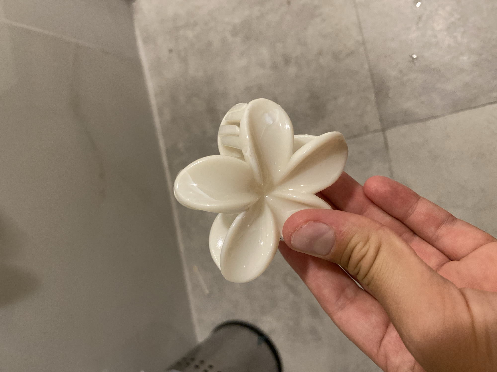 A person's hand holds a white, flower-shaped hair clip in front of a tiled wall and floor. The hair clip has five petals and a ribbed interior for gripping hair. The hand appears to be a right hand, with the thumb and index finger holding the clip. The background consists of light gray, rectangular tiles on the wall and floor, with some subtle variations in shading and texture. The overall scene suggests an indoor environment, possibly a bathroom or vanity area. The image does not provide any information about the city of Aksu, Turkey.