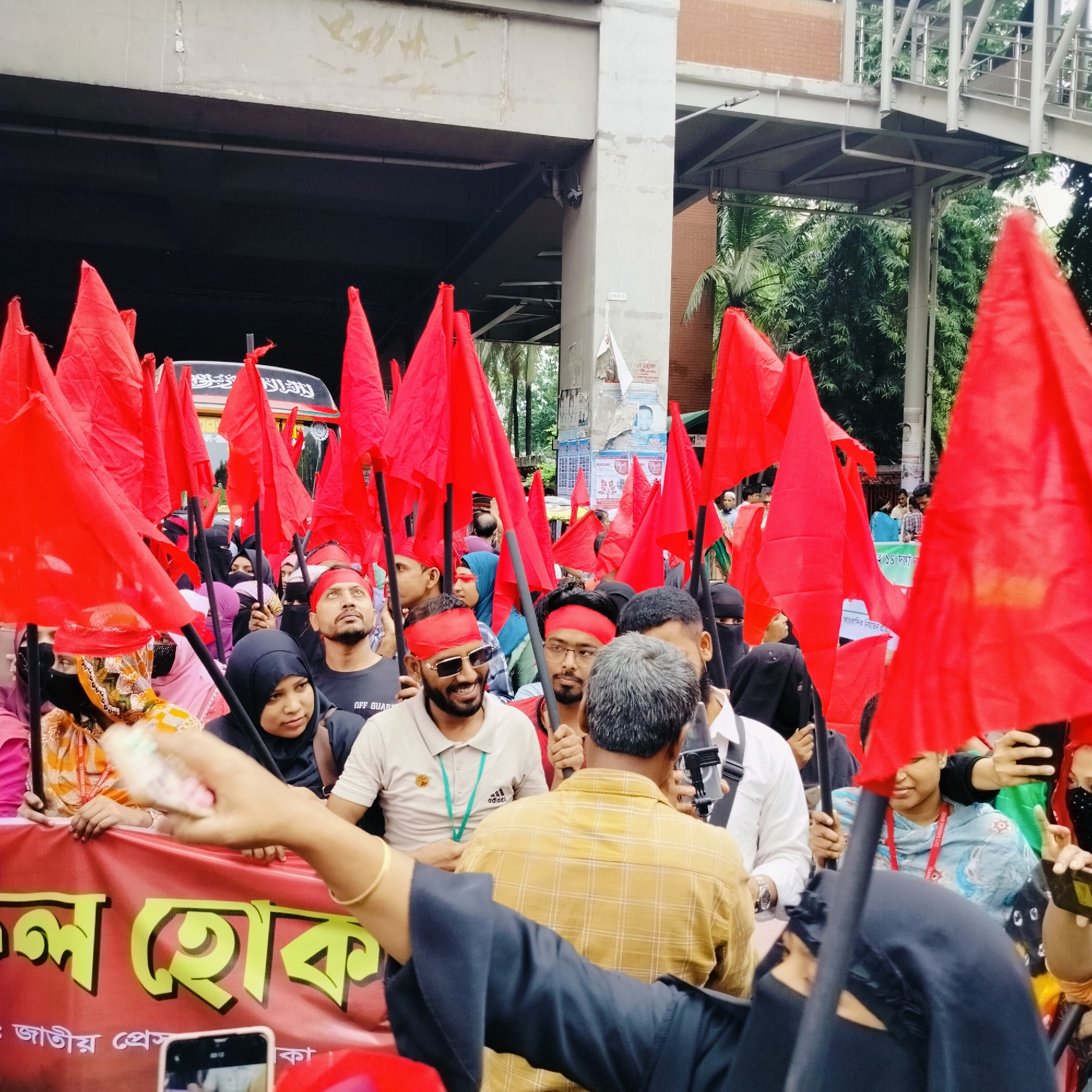 This media file captures a vibrant street protest or rally taking place in Dhaka, Bangladesh. A dense crowd of people, including both men and women, is visible, many carrying numerous red flags on poles, signifying their collective movement. A significant number of men wear red headbands, while several women are dressed in hijabs or niqabs, indicating diverse participation.
In the foreground, a prominent red banner with yellow Bengali text reads "ল হোক" (Lo Hok), which likely forms part of a larger slogan. Further text on the banner includes "জাতীয় প্রেস" (Jatiyo Press), suggesting a connection to the National Press Club, a known protest site in Dhaka, and phrases like "১৪ দফা" (14 Dafa - 14 points/demands) and "সাংবাদিক নির্যাতন প্রত" (Sangbadik Nirjaton Prot - Against Journalist Torture), clearly outlining the protest's objectives related to journalistic freedom and demands.
The setting is an urban environment, characterized by large concrete structures overhead, possibly a flyover or metro line, and lush green trees in the background. A white bus with some Arabic script is partially visible behind the flags. Participants appear engaged, some smiling while others look on with focused expressions, seemingly attentive to a speaker or a central activity. Details such as a person holding out what appears to be money or paper, and another recording with a smartphone, add to the dynamic nature of the event. The scene is illuminated by diffused daylight, indicative of an outdoor daytime gathering.