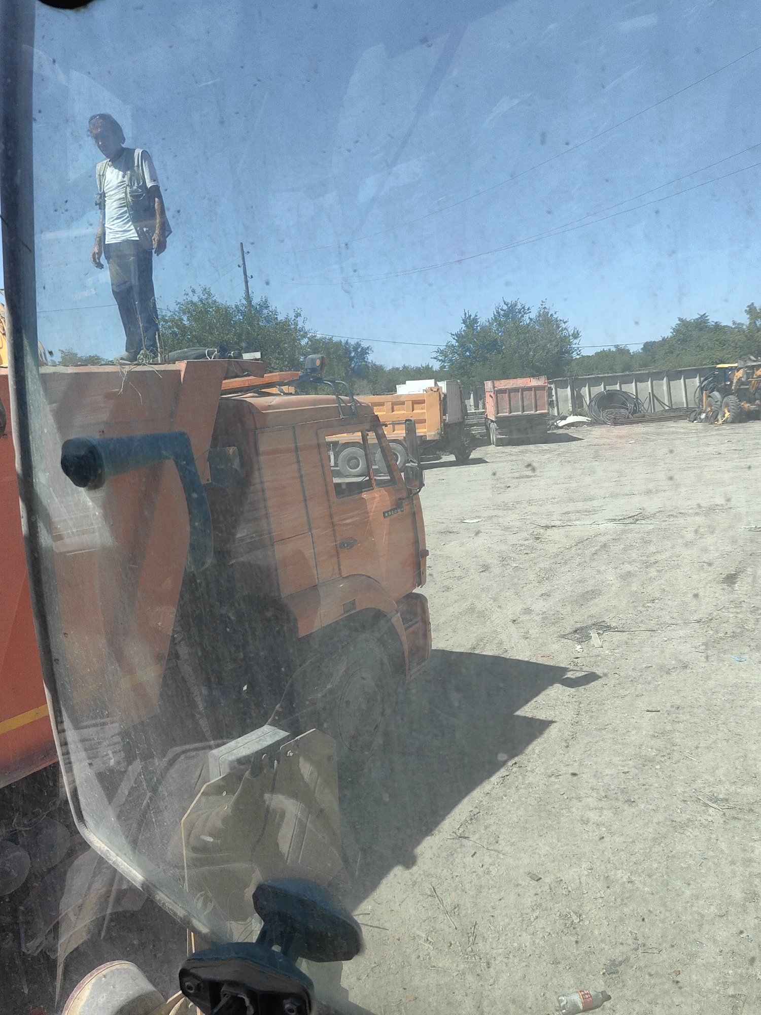 In Shymkent, Kazakhstan, a person stands atop an orange truck. The person, wearing a white t-shirt, a green vest, and dark pants, is positioned on the edge of the truck's load bed, looking down and to their left. The truck is parked on a dirt lot, with other construction vehicles and debris visible in the background under a clear blue sky.