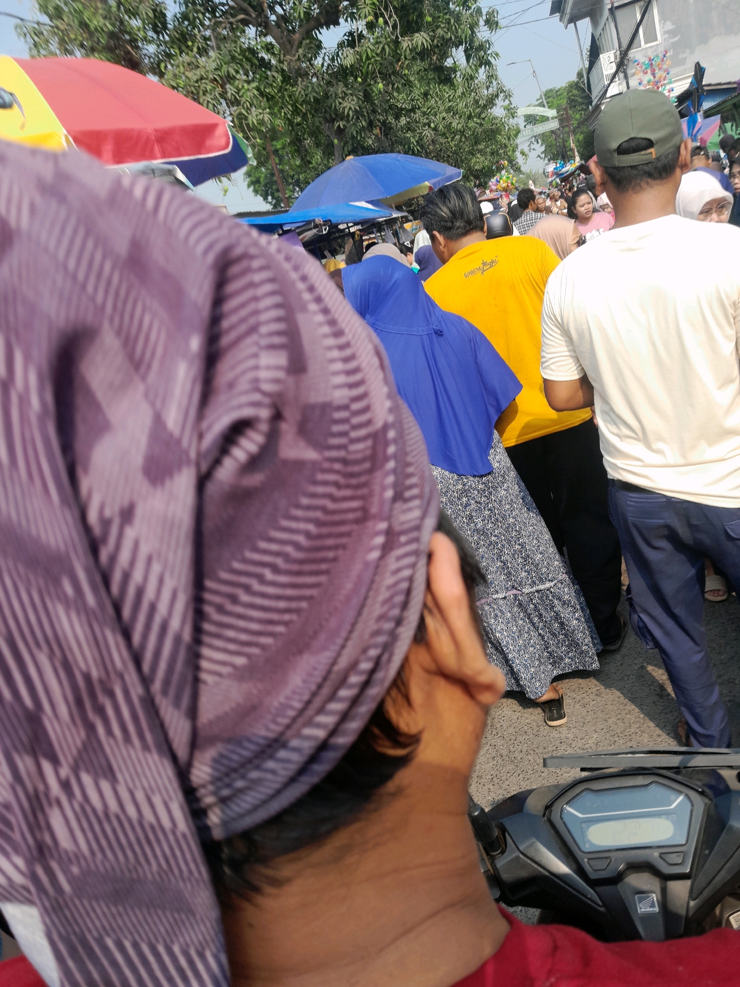 This is a medium shot, taken from a first-person perspective, of a person riding a motorcycle on a street in Jogosatru, Indonesia. The rider's head and shoulder are visible in the foreground, with a purple and white patterned headscarf or fabric covering their head and neck. The motorcycle's handlebars and dashboard are in the lower right corner.
In the middle ground, there are several people walking on the street, some under colorful umbrellas. A person in a blue hijab and a patterned dress is visible, as is a person in a yellow t-shirt and black pants. Another person in a white t-shirt and blue pants is standing to the right. Further back, more people and market stalls are visible, suggesting a busy street market or festival.
The background shows trees and buildings, indicating an outdoor urban environment. The lighting suggests it is daytime. The scene is crowded and appears to be a public gathering or event.