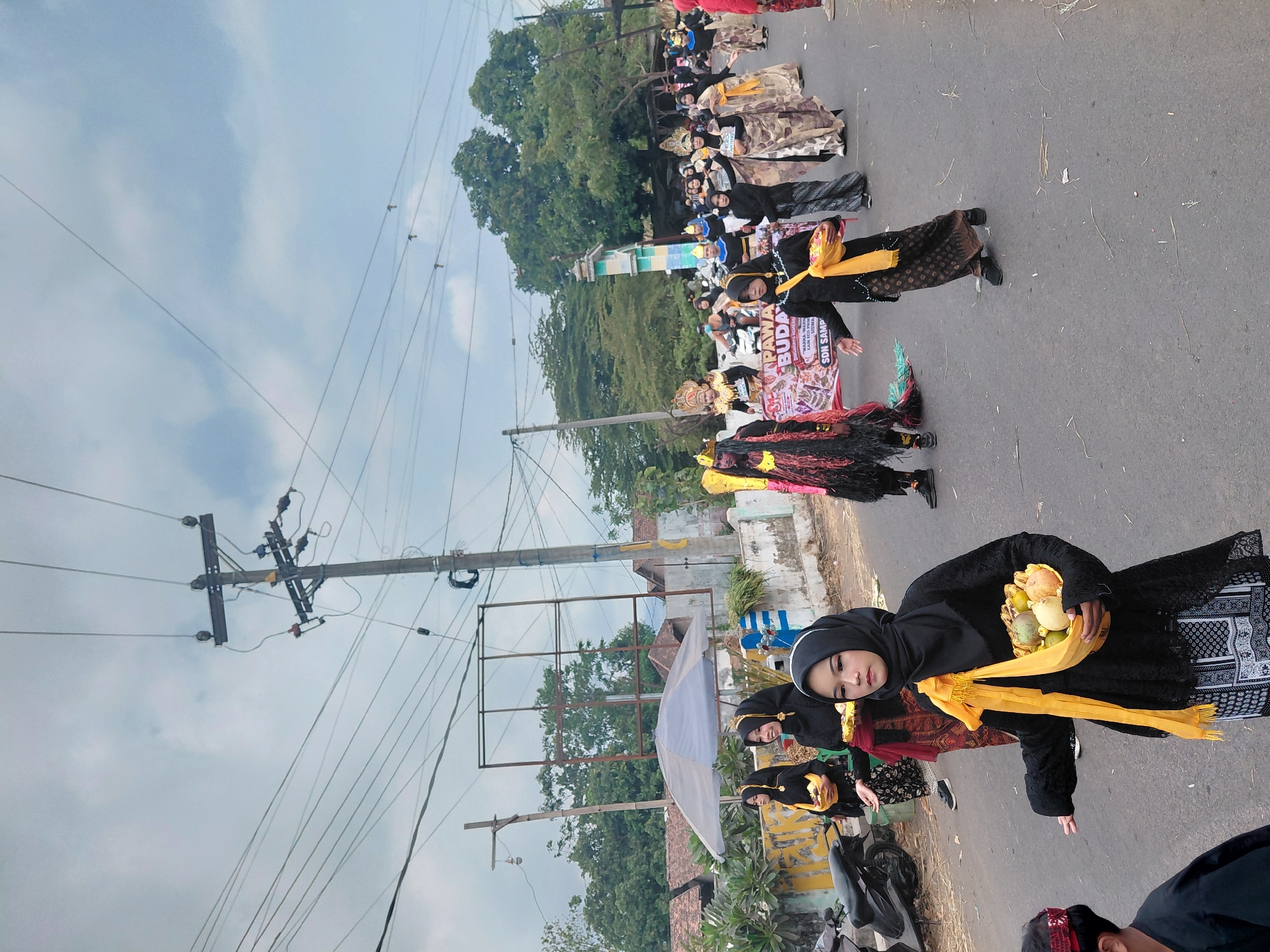 A parade is taking place on a street in Bimo, Indonesia. The street is paved and lined with houses and trees. The sky is cloudy.
Several people are participating in the parade, many of whom are dressed in traditional Indonesian attire. Some of the participants are wearing elaborate costumes, including a person in a black, fringed costume with a headdress. Others are dressed in black hijabs and long dresses, some carrying baskets of fruit. A banner with Indonesian text is visible in the background.
In the foreground, a young woman wearing a black hijab and dress is holding a basket of fruit. To her left, a motorcycle is parked on the side of the road. Power lines and utility poles are visible overhead. The overall scene depicts a cultural event or festival.