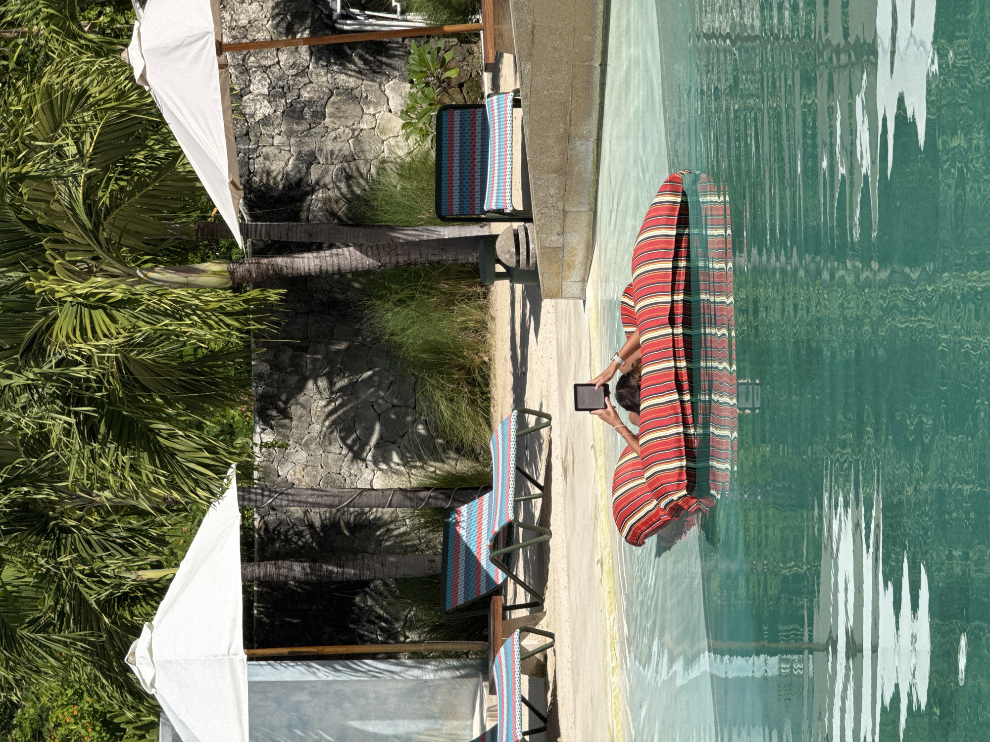 This outdoor, high-angle shot captures a serene poolside scene in a tropical setting, consistent with Nusa Penida, Indonesia. The main subject is a person relaxing in a swimming pool, partially submerged. They are wearing or wrapped in a vibrant red, white, and blue striped garment or towel and are engrossed in reading from a small, dark e-reader or tablet held in their hands.
The pool water is a clear, rich blue-green, displaying gentle ripples and reflecting the surrounding palm trees and sunlight. Adjacent to the pool, a light-colored paved area hosts two empty sun loungers, each with cushions matching the person's striped attire in red, white, and blue patterns.
The background is dominated by lush tropical vegetation, including large palm trees with broad fronds that cast strong shadows across a stone wall and the ground, indicating bright daylight, likely midday or early afternoon. Parts of white fabric umbrellas are visible, suggesting a resort or hotel environment. The overall ambiance is one of relaxation and leisure. No specific text is legible on the e-reader's screen.