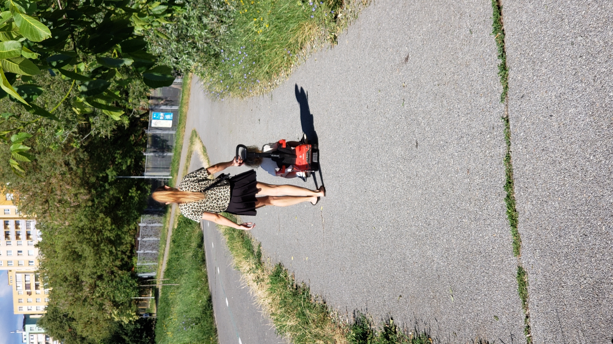 In Prešov, Slovakia, on a paved pathway, a woman with blonde hair pulled back in a ponytail, wearing a leopard print top and a black skirt, walks away from the camera. She is holding the handle of a red ride-on toy car, which is occupied by a young child. The child is wearing a white shirt. The pathway appears to be a bike or walking path, with a grassy area and trees to the left, and more vegetation to the right. In the background, there are residential buildings and a chain-link fence. The lighting suggests it is daytime.
