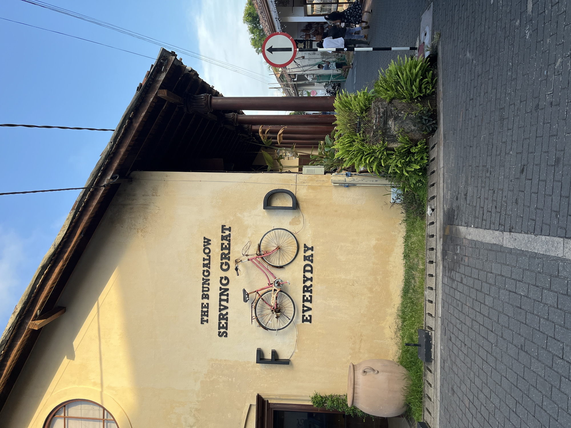 A pale yellow stucco building with a weathered facade stands on the left side of a cobblestone street in Galle, Sri Lanka. The building features a dark wooden roof with decorative eaves and columns. Black cutout letters on the facade spell out "THE BUNGALOW SERVING GREAT FOOD EVERYDAY." A pink vintage bicycle is mounted on the wall, serving as a decorative element. A large, light brown ceramic pot sits on the grass in front of the building, to the left of the entrance. Green ferns and other vegetation grow around a stone structure to the right of the building, near the street. The street continues into the distance, lined with more buildings and people walking. A white and black striped pole with a circular sign showing a black arrow pointing upwards is positioned on the right side of the street. The sky is blue with a few scattered clouds. The scene appears to be set in a commercial or tourist area.