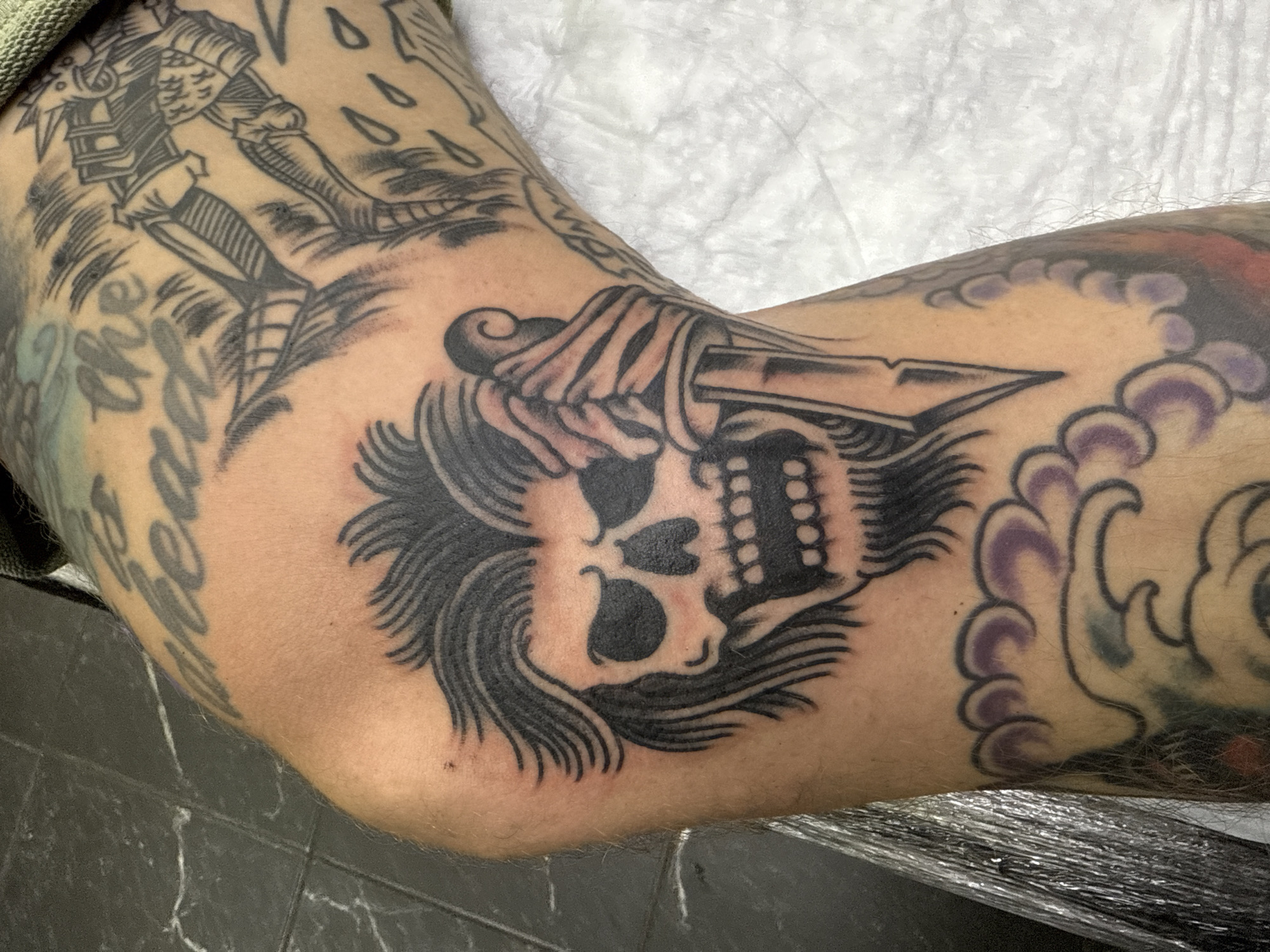 A close-up shot captures a heavily tattooed human arm, likely taken indoors in a professional setting such as a tattoo studio in Moscow, Russia. The primary focus is a recently completed tattoo located on the inner elbow area, featuring a stylized skull with bold black eyes, nose, and teeth. Wavy, dark hair or flame-like elements frame the skull, and a skeletal hand is depicted holding a dagger that pierces the top of the skull. The skin immediately surrounding this central tattoo shows noticeable redness, indicating its freshness.
Portions of other tattoos are visible on the arm. On the upper left, part of a larger black-and-grey tattoo depicts a standing figure, possibly a warrior or knight. Below this, faint black script lettering is present, but it is largely obscured and difficult to read clearly. On the right side of the arm, towards the forearm, there are sections of another tattoo with wavy, cloud-like patterns outlined in black, some of which are shaded in light purple and grey.
The overall style of the visible tattoos suggests a traditional or neo-traditional aesthetic, characterized by strong black outlines and solid ink fills. The background consists of a dark, possibly tiled floor on the bottom left, and a white, textured surface—perhaps a protective sheet or towel—on the upper right. A piece of reflective plastic wrap is visible in the bottom right corner.
