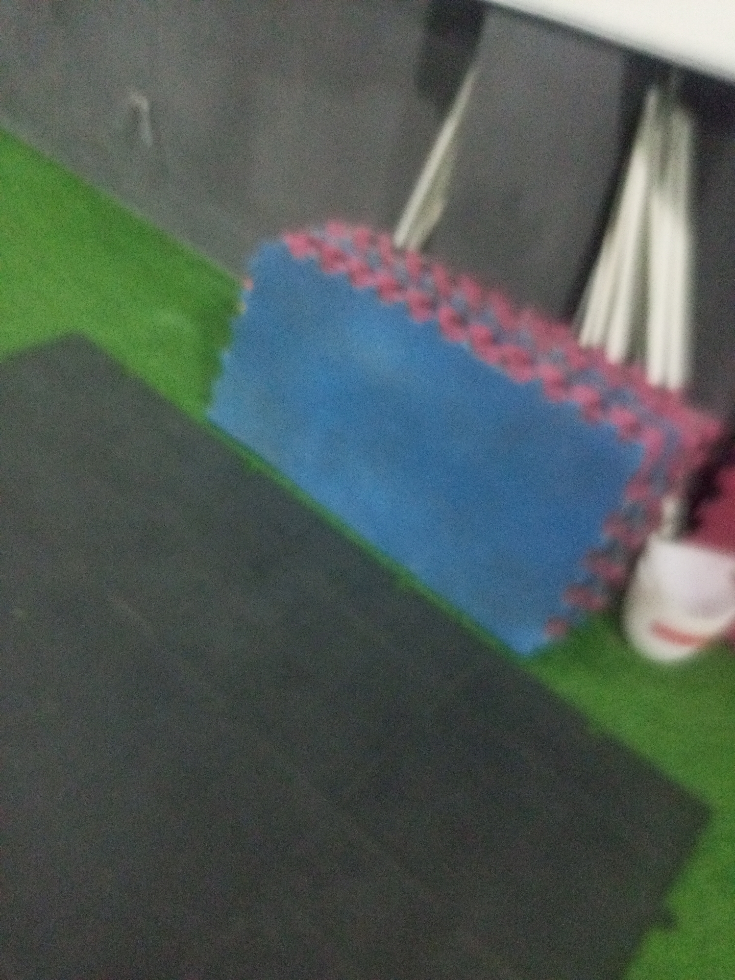 The image shows a close-up, out-of-focus view of interlocking foam mats. A large, blue mat with pink edges is positioned diagonally on top of a dark gray mat. The mats are on a green surface, which may be artificial turf. In the background, there are blurred vertical white and gray elements, possibly part of a structure or equipment. A white object with red accents, possibly a shoe, is partially visible on the right side of the image. The scene appears to be an indoor or covered area. There is no discernible context to place it within Juan Griego, Venezuela.