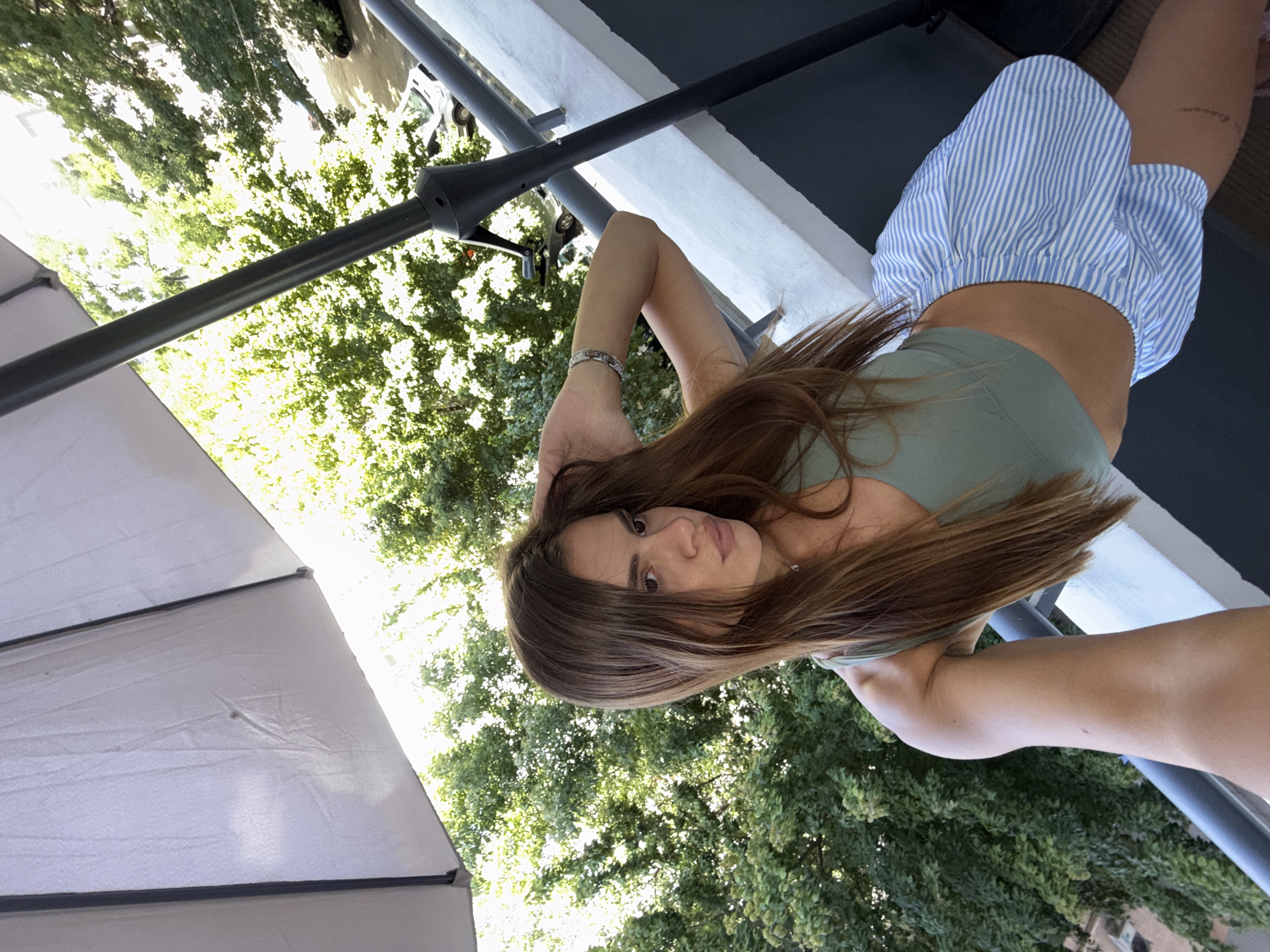 A young woman with long brown hair is posing on a balcony in Plovdiv, Bulgaria. She is wearing a green crop top and blue and white striped shorts. She is holding her hand to her hair and looking towards the camera. In the background, there are trees and buildings, and a large umbrella is visible in the foreground.