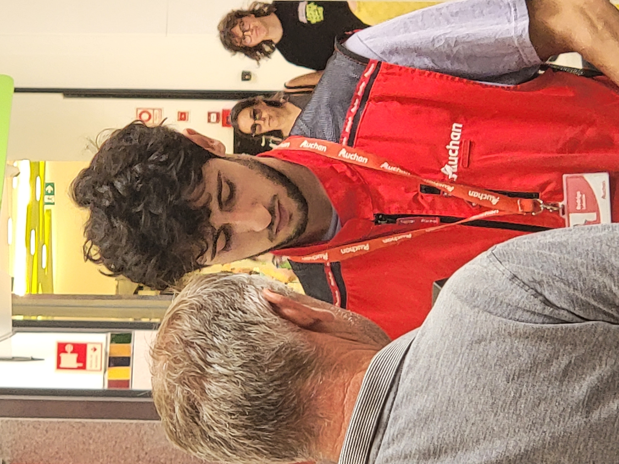 Two individuals are the primary focus of the image. The person in the foreground, on the left, is an older male with gray hair, seen from the back. He is wearing a gray t-shirt with a striped collar. Directly in front of him, slightly to the right, is a younger male with curly dark hair. He appears to be an employee, wearing a red vest over a light purple t-shirt. The red vest has the "Auchan" logo and several lanyards with ID badges attached. The background shows the interior of a brightly lit store, likely a supermarket. To the right of the younger male, a woman with dark hair and sunglasses is visible, and further to the right, a person with curly hair and glasses is also present. The environment appears to be a retail space with merchandise racks and signage visible in the background. The store is likely Auchan, a hypermarket chain, and given the context of Faro, Portugal, this scene is situated within such a store in that city.