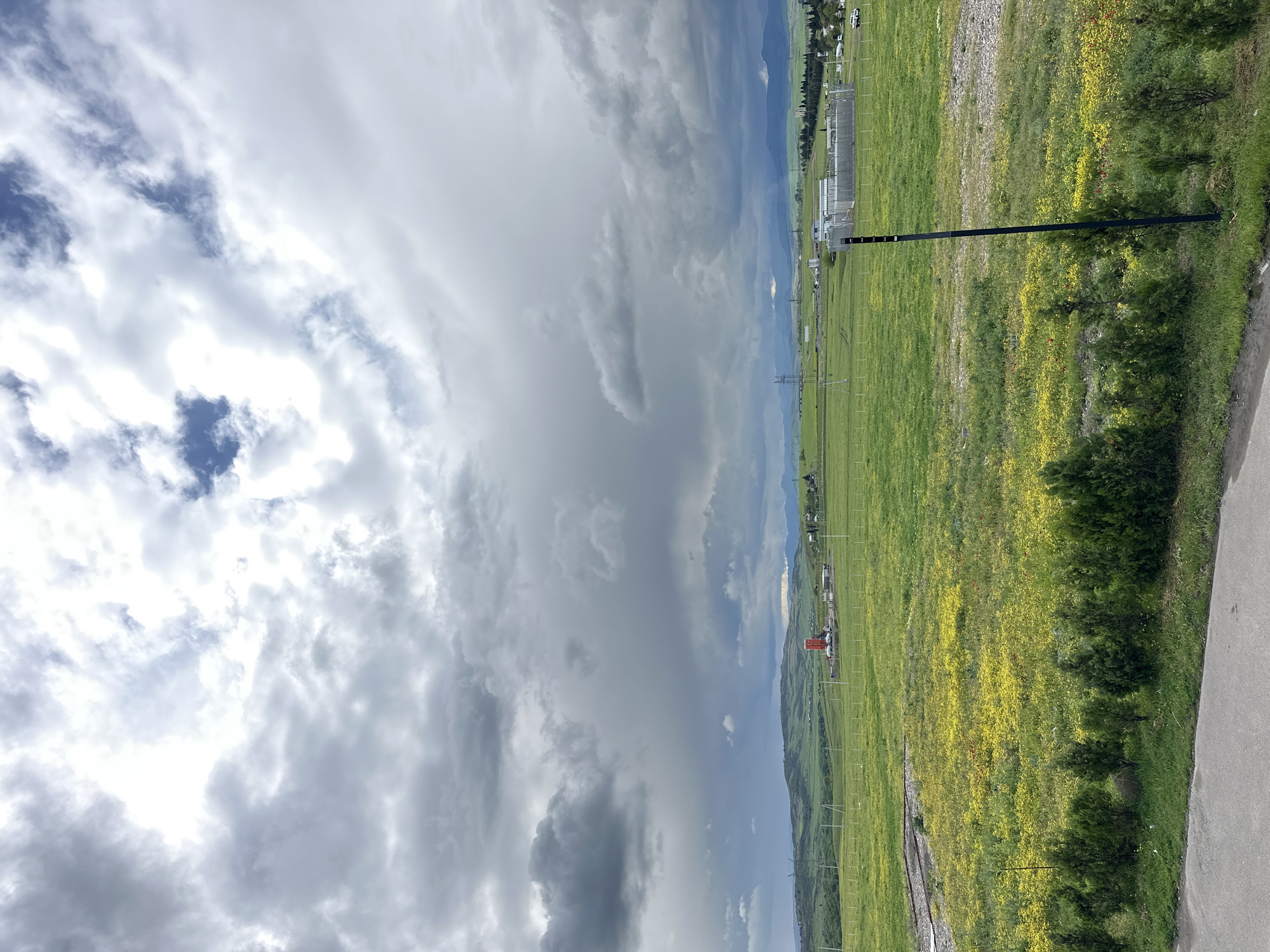 The image depicts a landscape with a cloudy sky covering approximately half of the frame. Below the sky, a green field stretches out with patches of yellow flowers. A black pole stands in the middle ground. In the background, there are buildings and infrastructure, possibly indicating an industrial or urban area. The location appears to be near Marneuli, Georgia, suggested by the hilly terrain in the distance.