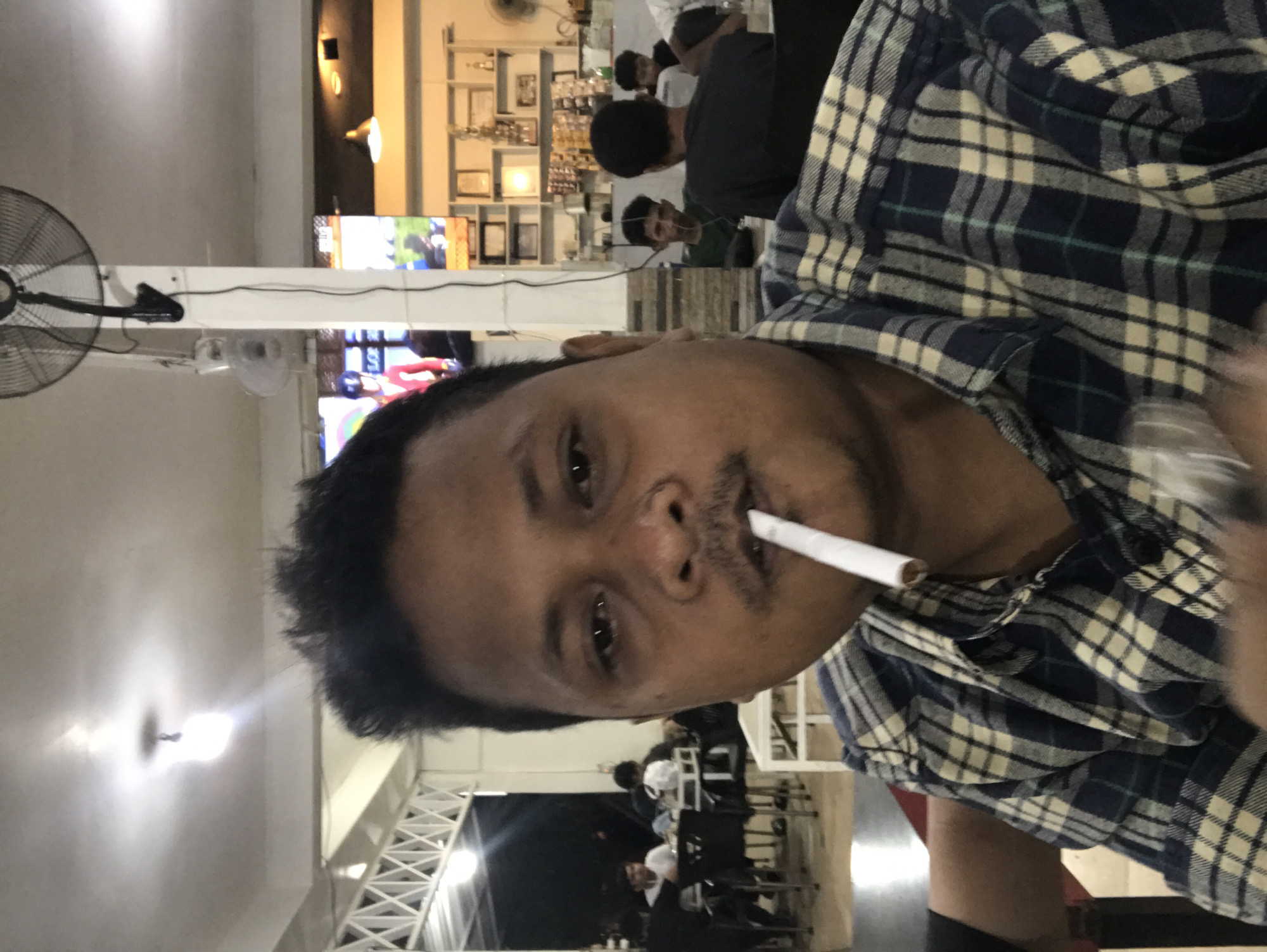 The image shows a man indoors, holding a cigarette in his mouth. He is wearing a blue and white checkered shirt. In the background, there are tables and chairs, suggesting a cafe or restaurant. Other people are visible in the background. The environment appears to be a casual dining establishment, possibly in Lhokseumawe, Indonesia. There are TV screens displaying content, and ceiling fans are present.