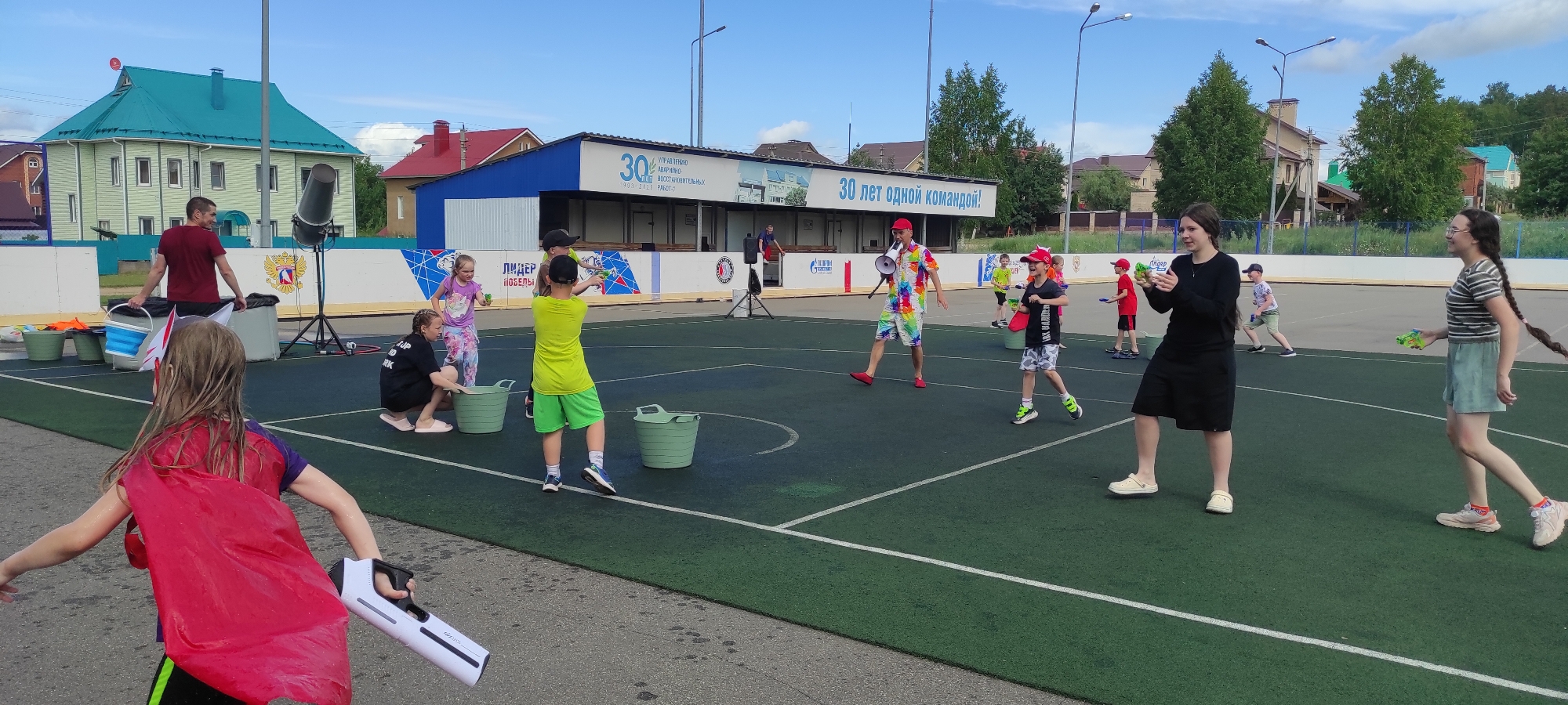 The image depicts an outdoor summer scene on a green athletic field, likely in Noviy, Russia, with a building in the background. The field is marked with white lines, and several people are engaged in a water-based activity.
In the foreground, a young person wearing a red cape is holding a white water gun. To their right, a woman in a black dress and white clogs is holding a green water gun. Further to the right, another young person with a long braid, wearing a striped t-shirt and a green skirt, is also holding a green water gun.
In the middle ground, a person in a colorful shirt and shorts, wearing a red hat, is holding a megaphone and appears to be leading the activity. Several children are scattered across the field, some holding water guns and buckets. One child in a bright yellow shirt and green shorts is aiming a water gun. Another child in a black shirt and patterned shorts is interacting with a bucket. Two younger children, one in a pink shirt and patterned leggings, and another in a red shirt, are also present.
In the background, a building with blue and white paneling is visible, featuring banners with Russian text. To the left of the building, a tall structure resembling a speaker or a fan is present. Houses with red and teal roofs are also visible in the distance, along with trees. The sky is partly cloudy. The overall atmosphere suggests a playful, organized event, possibly a children's party or a community gathering.