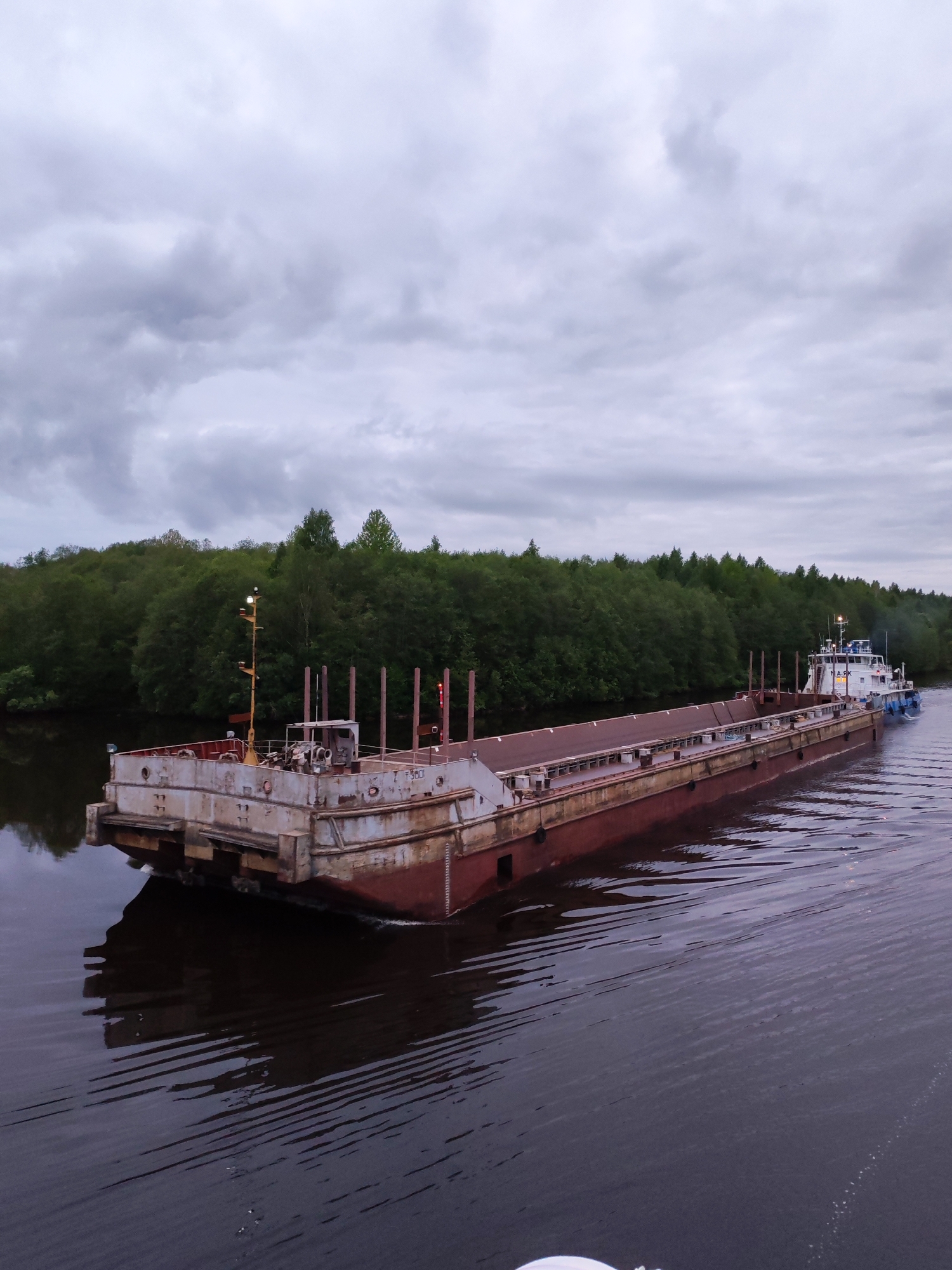 A long, weathered barge is being towed on a waterway in Volokov Most, Russia. The barge is rusty and appears to be made of metal. It has a low profile and is likely used for transporting materials or goods. The towboat is visible in the distance, pulling the barge along the water. The sky is overcast, casting a dim light on the scene. Trees line the banks of the waterway, providing a natural backdrop. The water is dark and still, reflecting the barge and the surrounding environment.