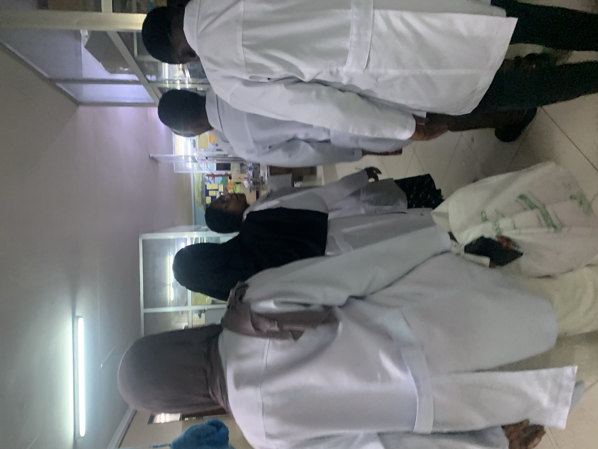 The image depicts a group of individuals in what appears to be a medical or laboratory setting. The scene is indoors, possibly in Dar es-Salaam, Tanzania.
Several individuals are wearing white lab coats, suggesting they may be healthcare professionals or students. One person is wearing a hijab. The individuals are standing, interacting, or observing something, though the specific action is unclear. There are also white linens.
The environment includes fluorescent lighting. The details provide a limited overview of the scene, which appears to be focused on a medical or educational context within an indoor setting.